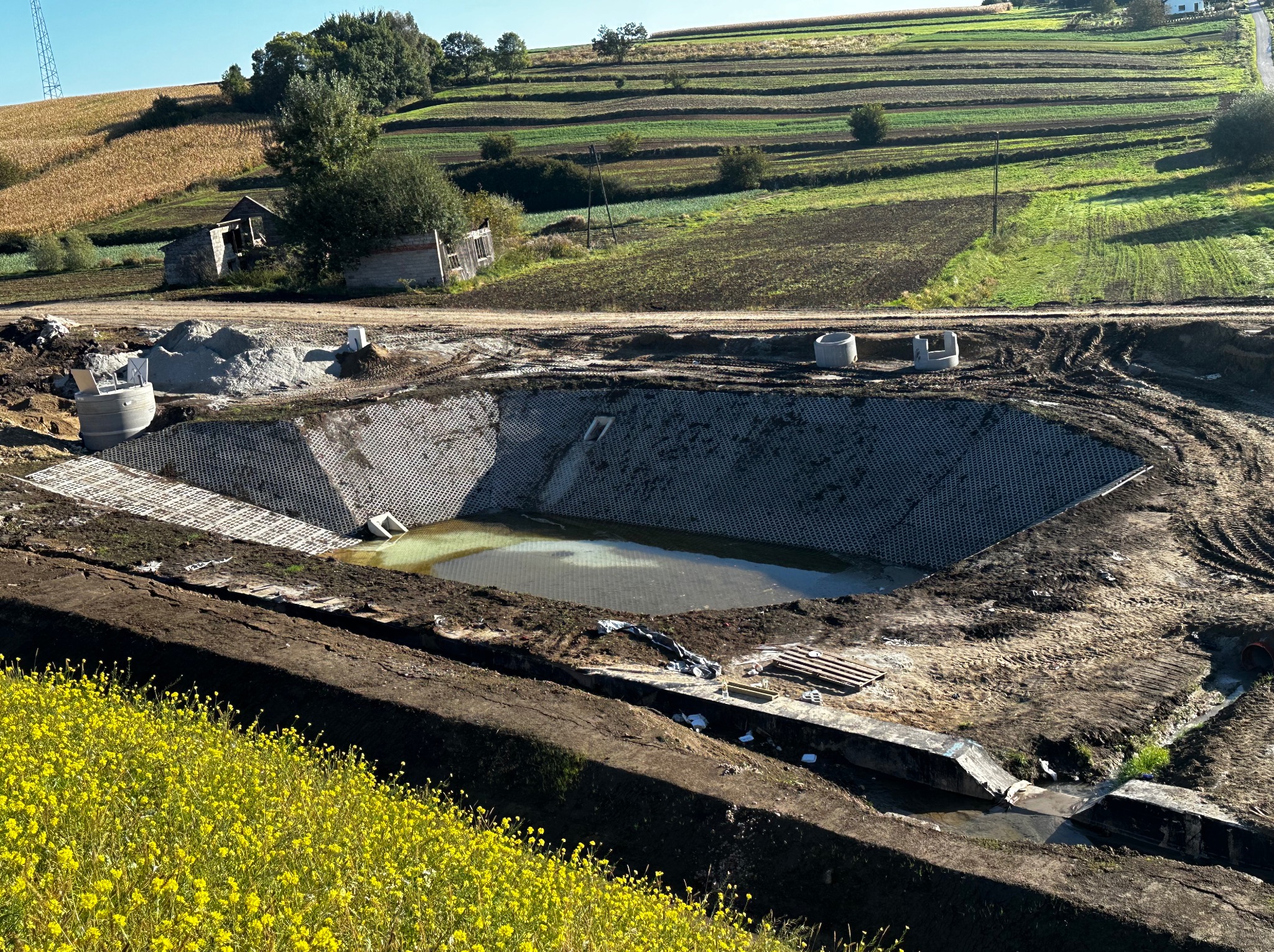 Budowa zbiornika retencyjnego na tle pagórkowatego krajobrazu. Ukończona betonowa konstrukcja z wodą w środku, widoczne elementy kanalizacji i żółte kwiaty.