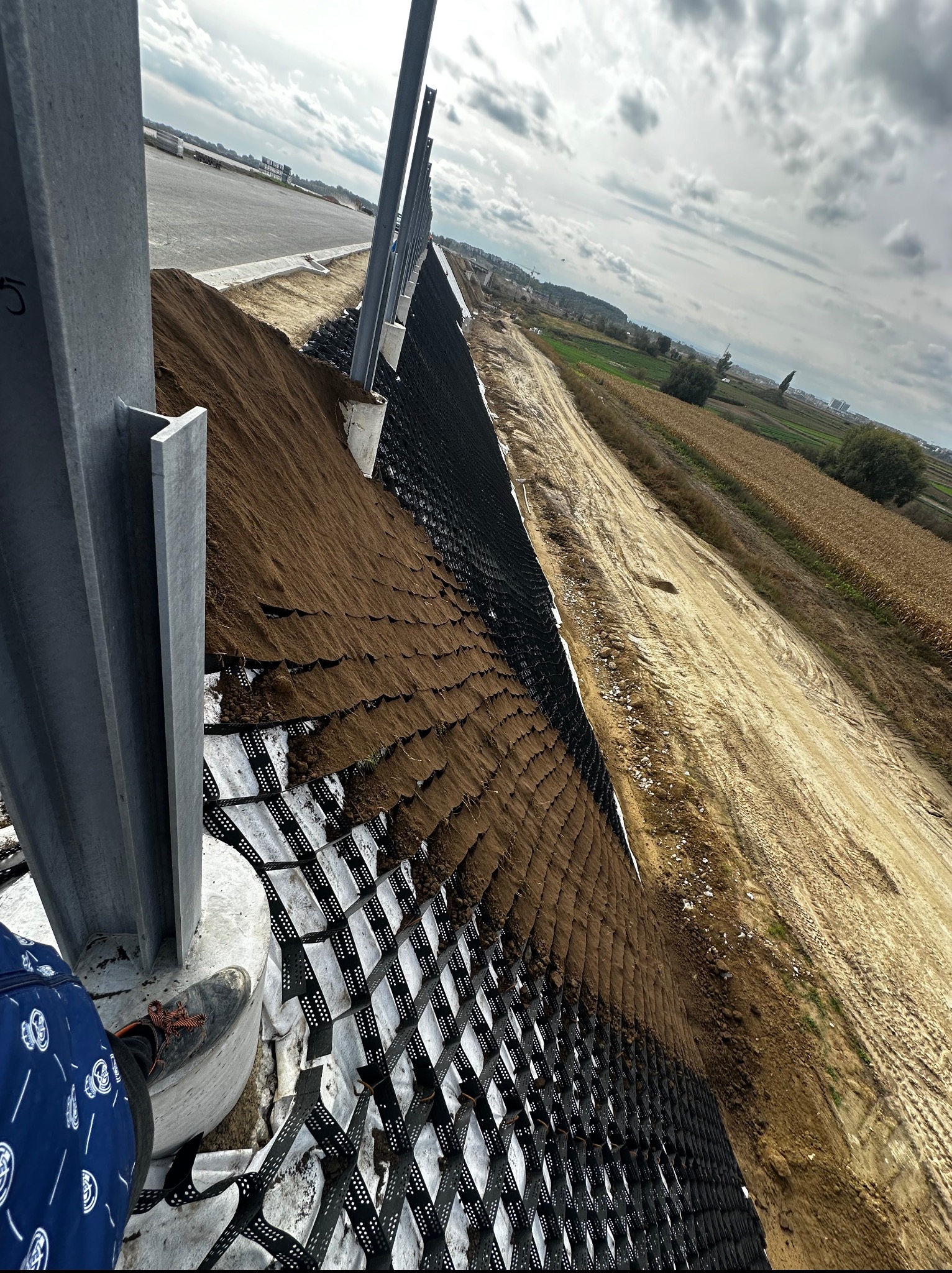 Ukończona stabilizacja skarpy z użyciem geokraty i palików, zabezpieczona ziemią. Widok na krajobraz rolniczy w tle, pochmurne niebo. Kąt ujęcia z dołu.