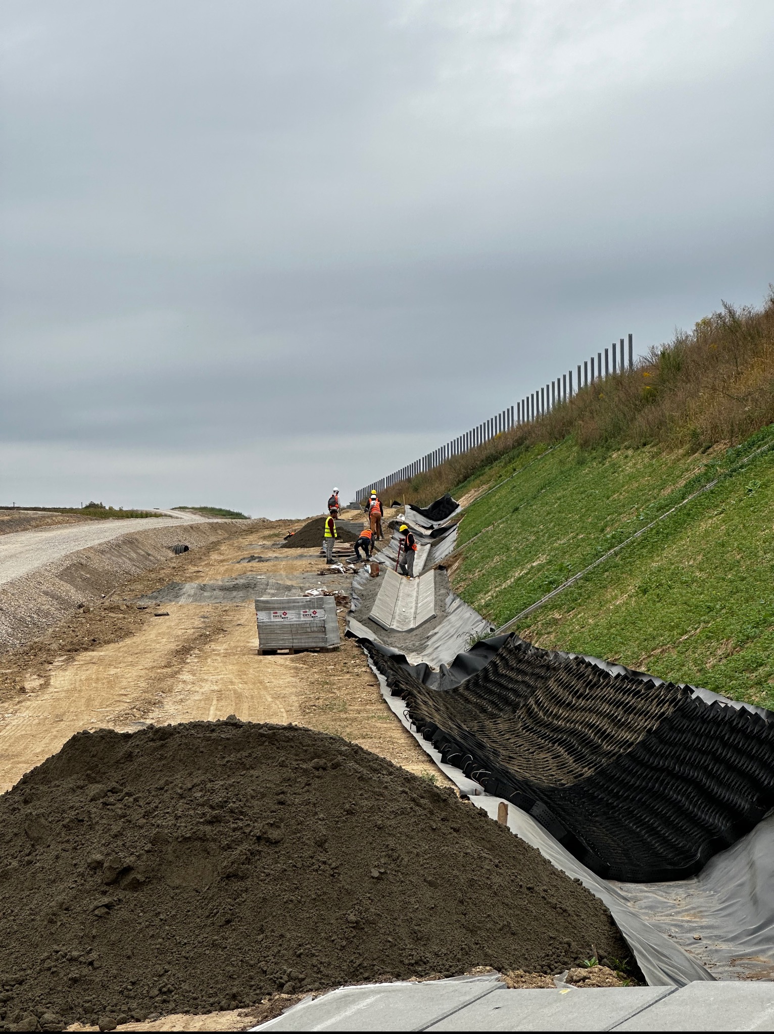 Budowa rowu melioracyjnego: robotnicy w odblaskowych kamizelkach układają betonowe elementy wzdłuż zbocza, zabezpieczonego geowłókniną i palisadą.