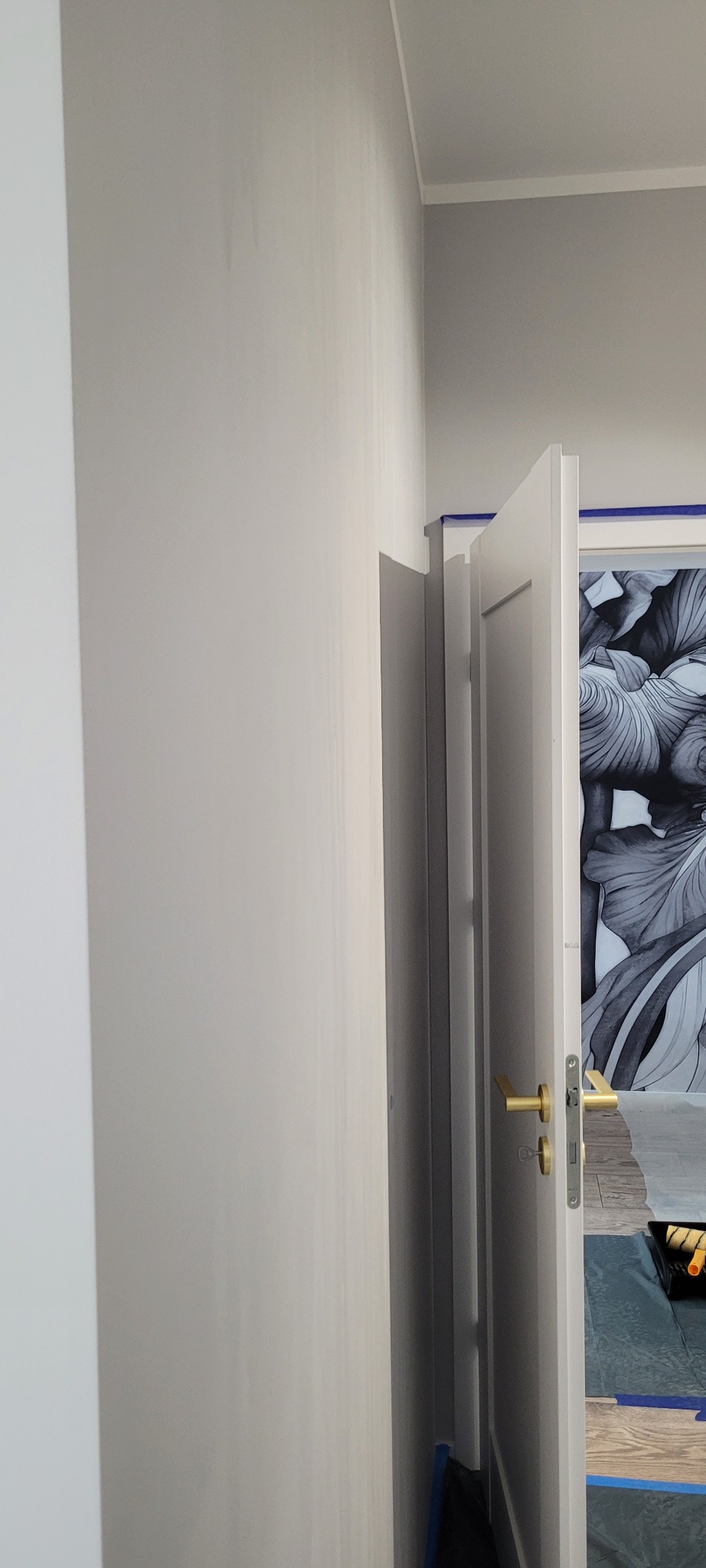 Świeżo pomalowana ściana w odcieniach szarości z otwartymi białymi drzwiami, zabezpieczonymi taśmą malarską. Widoczne narzędzia malarskie w tle.