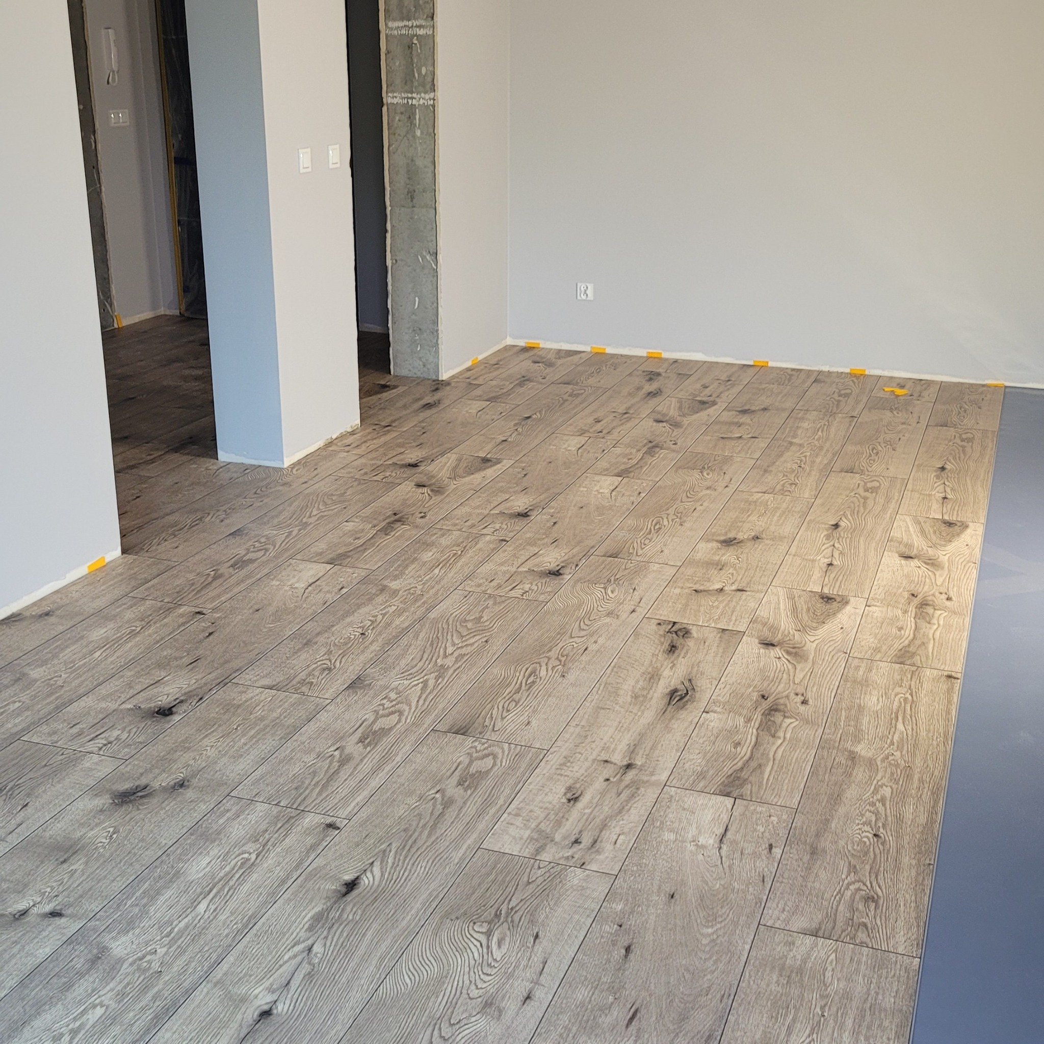 Ułożona podłoga z paneli drewnopodobnych w jasnych odcieniach szarości, z widocznymi elementami dystansowymi przy ścianach. W tle surowy betonowy filar.