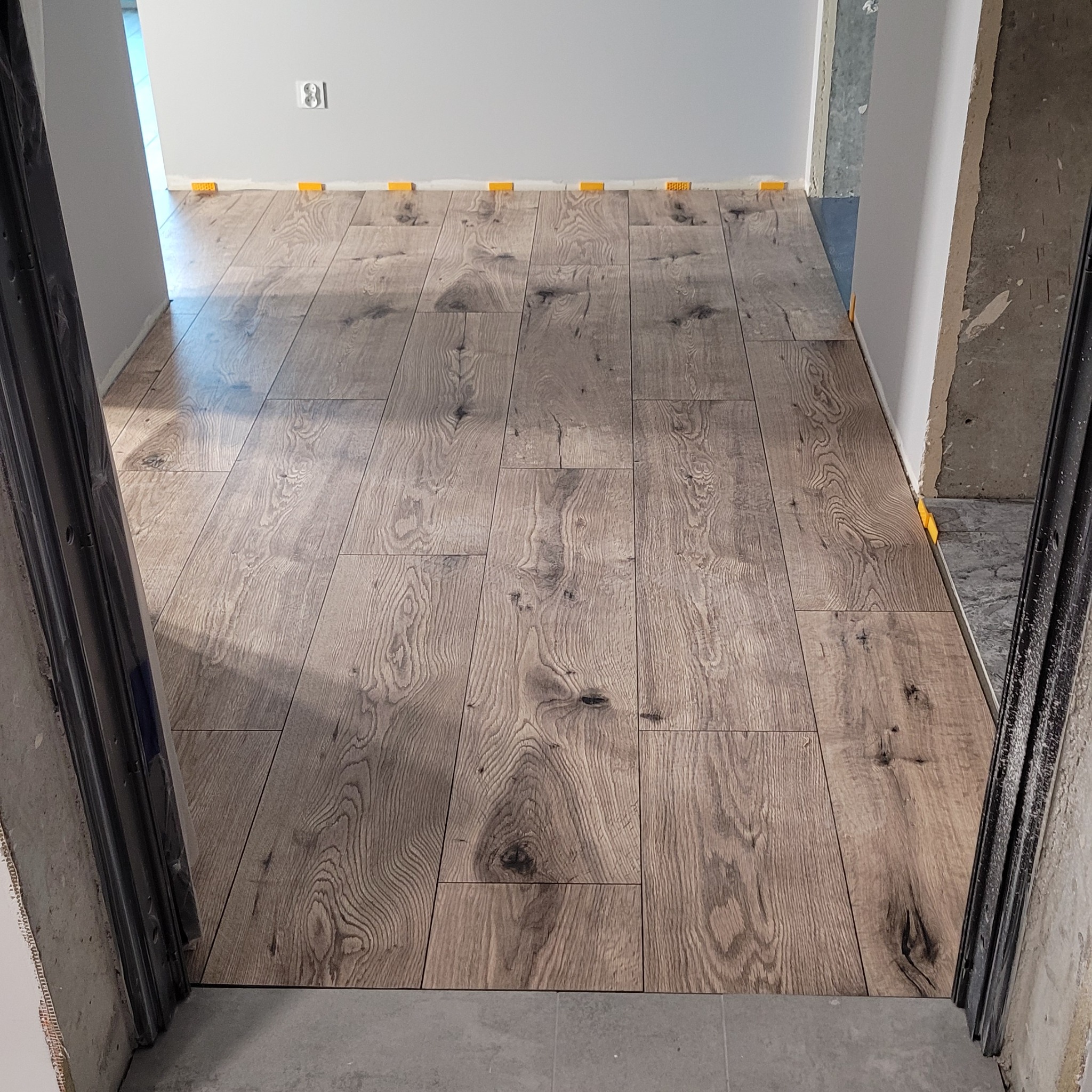 Ułożona podłoga z paneli w odcieniach brązu, widok z progu drzwi, z użyciem żółtych klinów dystansowych. Prace wykończeniowe w mieszkaniu w Poznaniu.