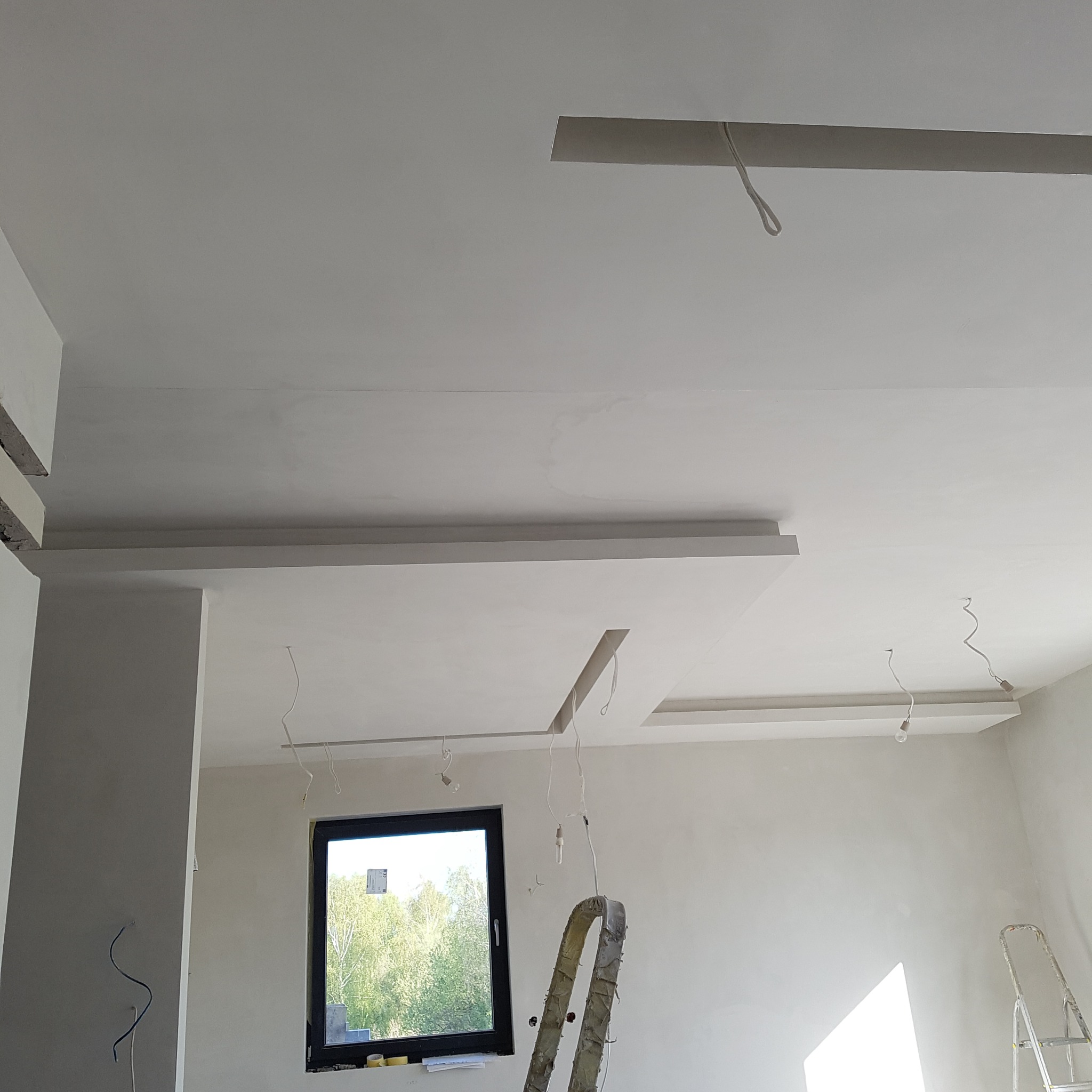 Białe, wykończone sufity podwieszane z oświetleniem w nowym, jasnym wnętrzu. Widoczne okno i drabina. Minimalistyczny design.