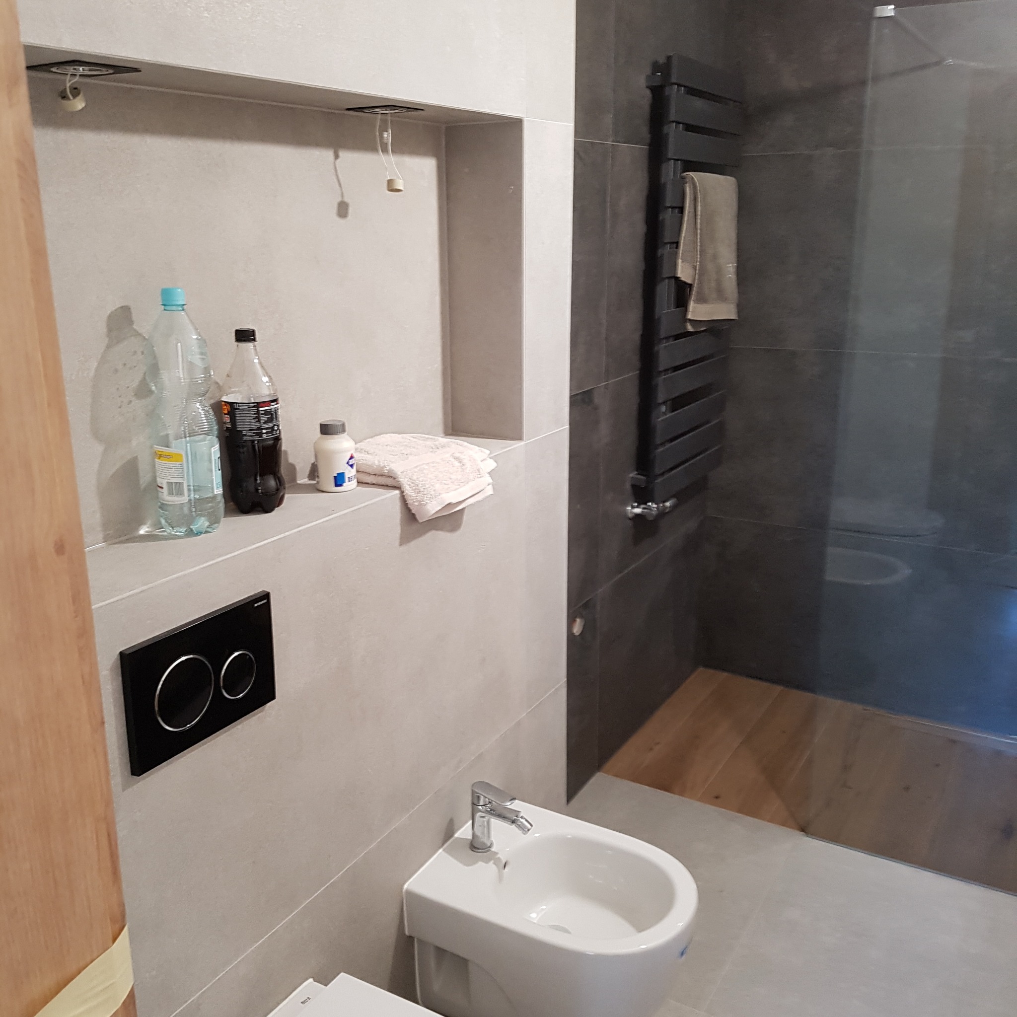 Nowoczesna łazienka z szarą glazurą, białym bidetem i grzejnikiem drabinkowym z ręcznikiem. Wnęka z oświetleniem i akcesoriami dodaje charakteru.