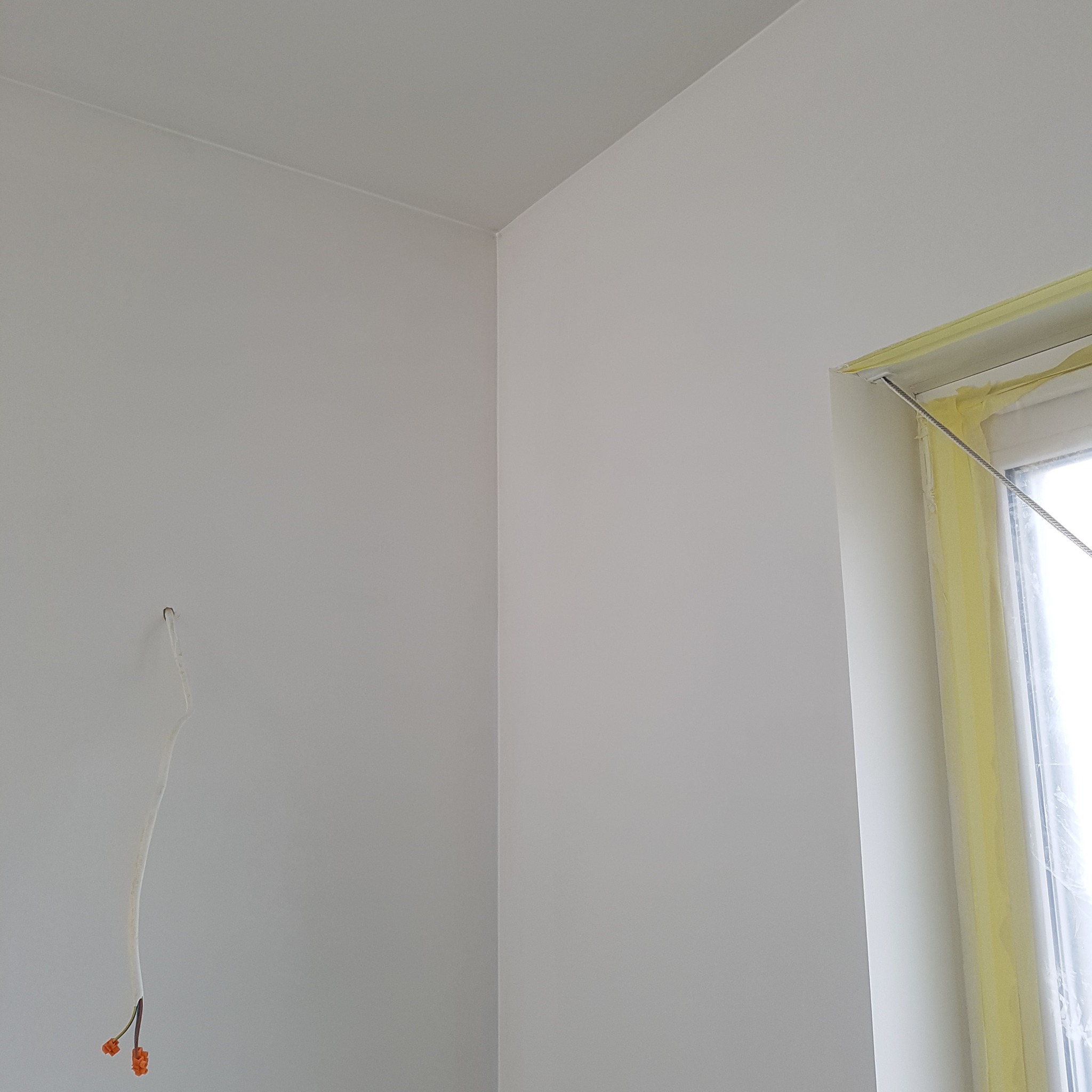 Świeżo pomalowane na biało narożniki ścian i okno zabezpieczone taśmą malarską. Z sufitu zwisa kabel elektryczny. Wykończenie wnętrz w minimalistycznym stylu.