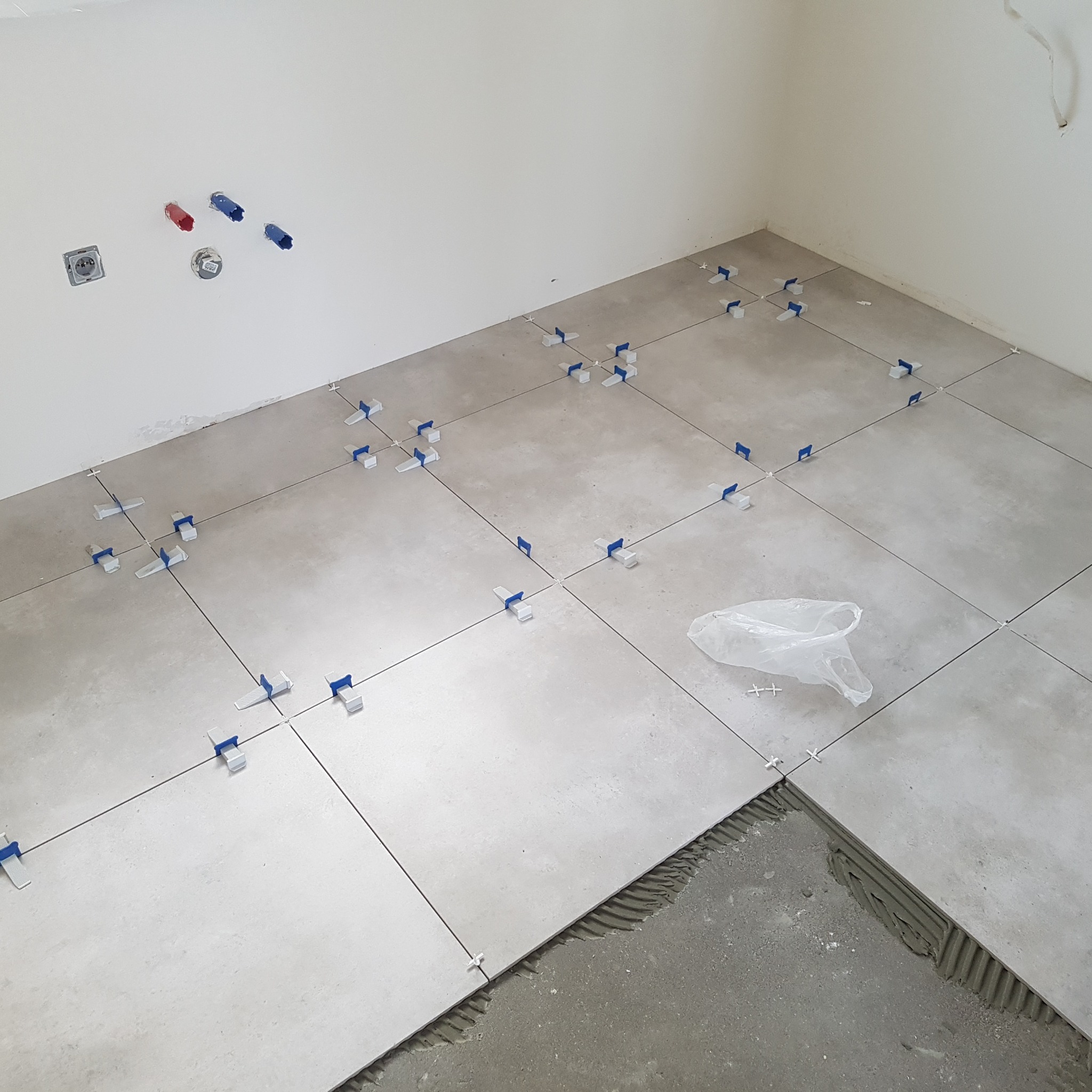 Jasnoszare płytki podłogowe w trakcie układania, z niebieskimi krzyżykami dystansowymi, widoczne instalacje wodne na ścianie. Częściowo ułożona podłoga.