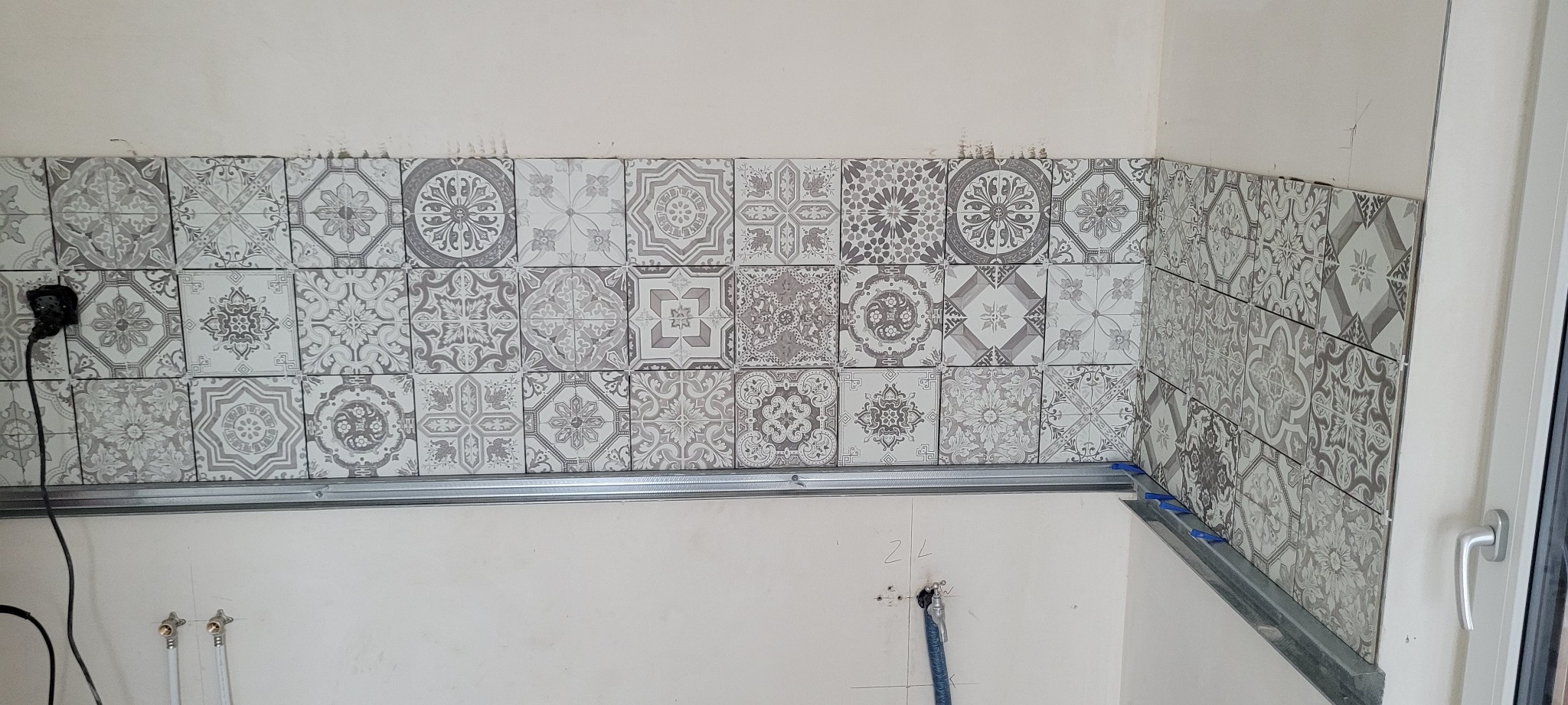 Układanie wzorzystych płytek na ścianie, widoczna listwa montażowa i krzyżyki dystansowe. Etap prac wykończeniowych w pomieszczeniu.