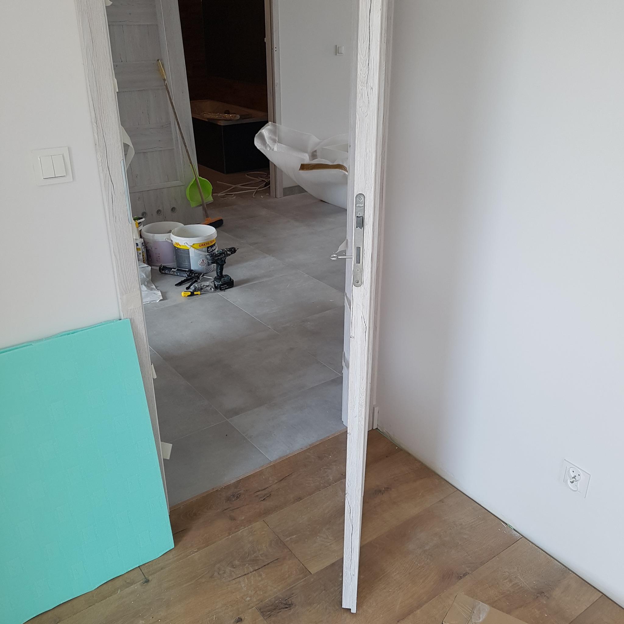Remont wnętrza: widok przez uchylone drzwi na podłogę z płytek i panele. Widoczne narzędzia, puszki z farbą i płyta izolacyjna. Świeżo pomalowane ściany.