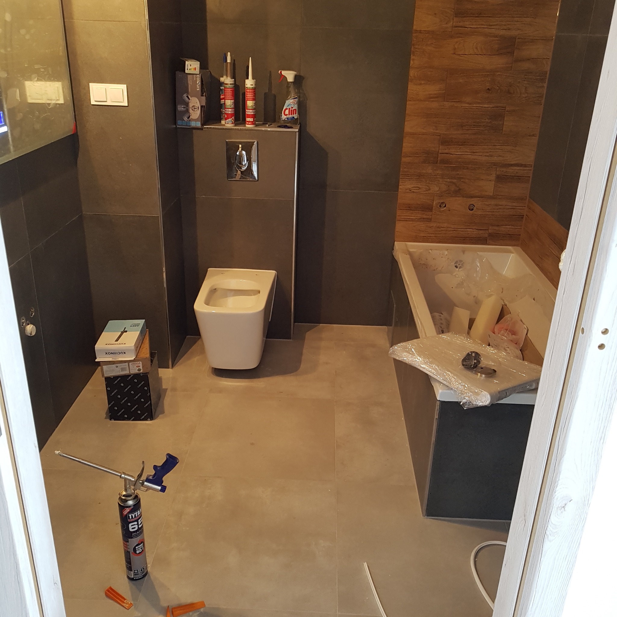 Wnętrze łazienki w trakcie prac wykończeniowych: zamontowana toaleta, wanna w folii, kartony i piana montażowa. Widoczne szare płytki i fragment drewnopodobnej okładziny.