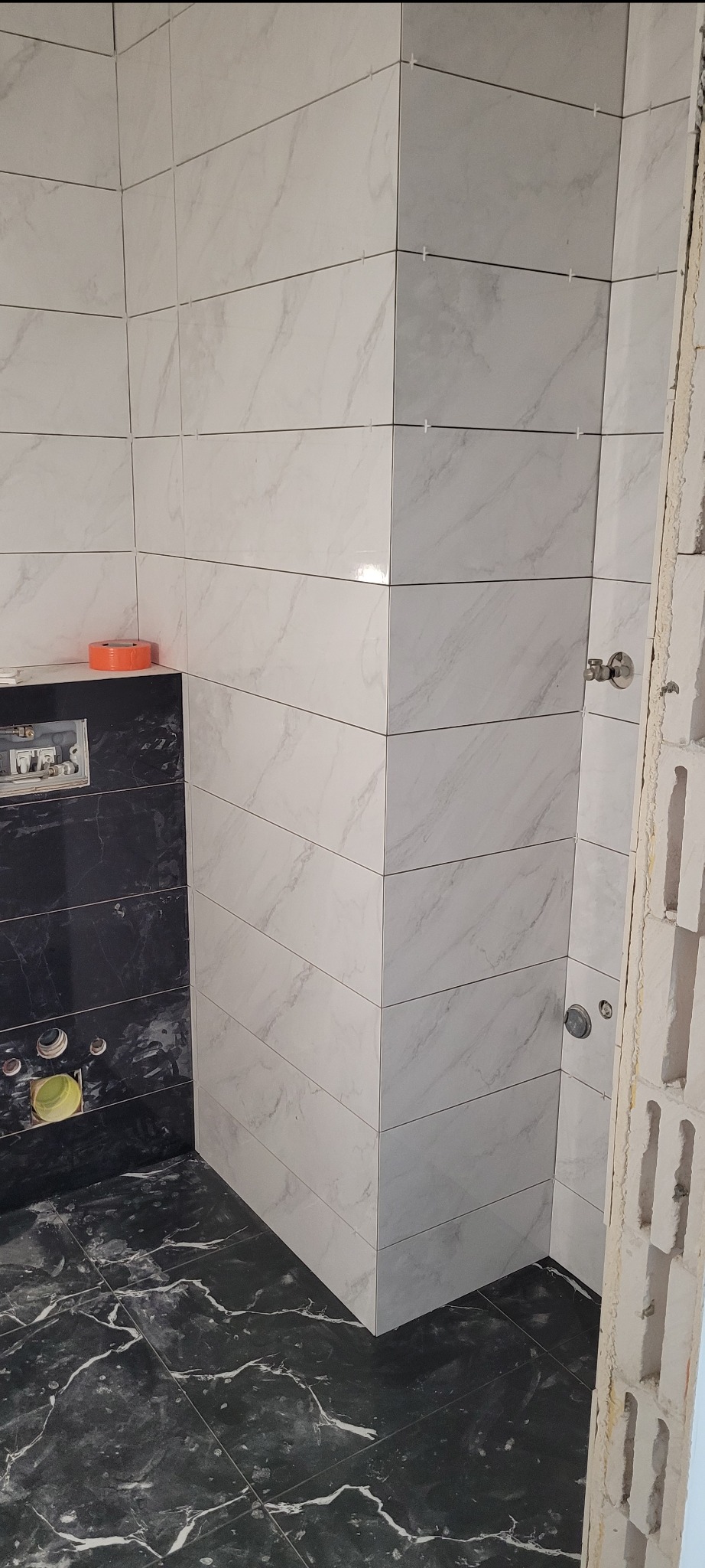 Wykończona łazienka z czarno-białymi płytkami imitującymi marmur. Widoczne krzyżyki dystansowe między płytkami na ścianie. Fragment ściany z pustaków.