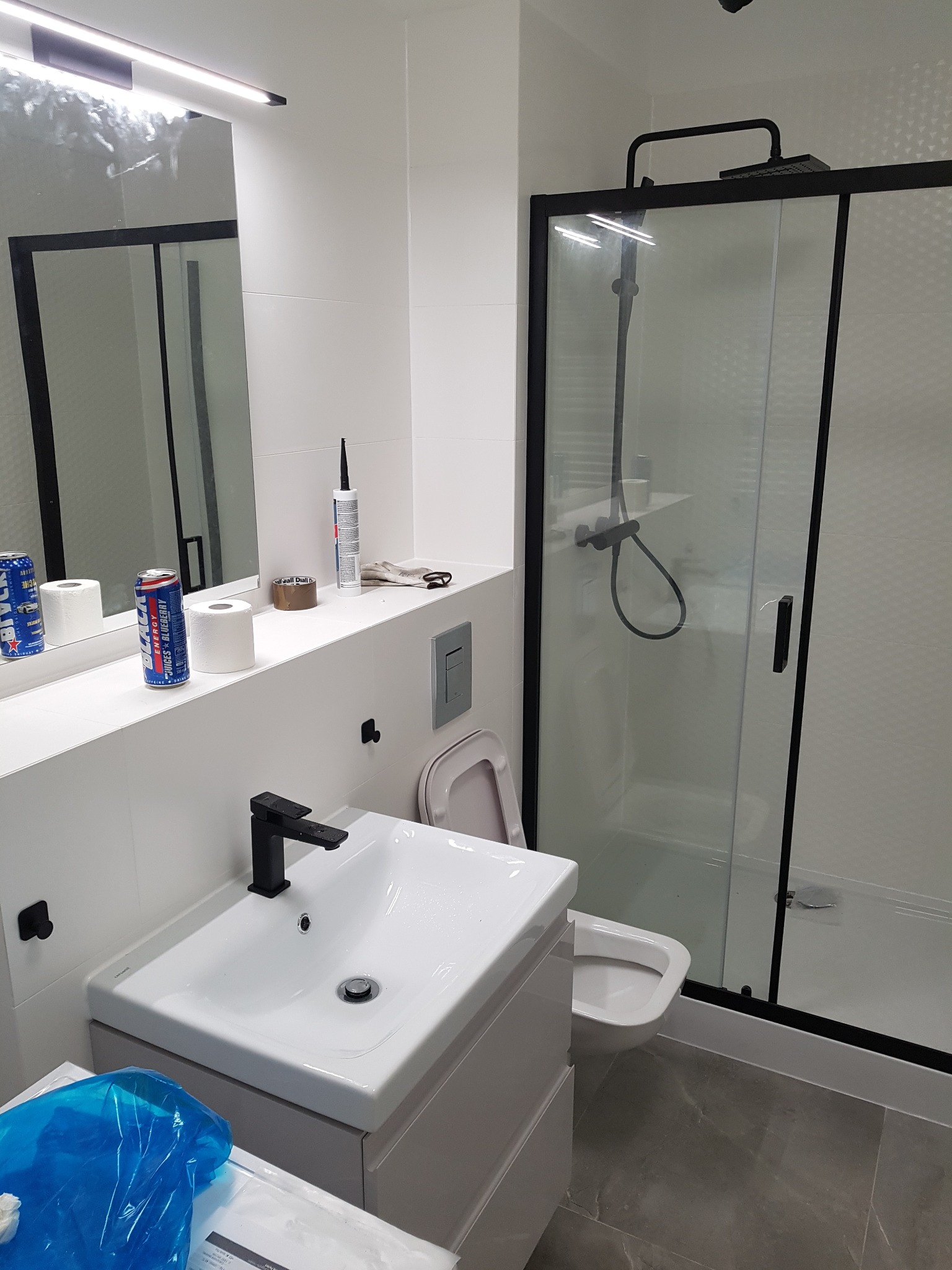 Nowoczesna łazienka po remoncie: białe płytki, czarna armatura, kabina prysznicowa z przesuwnymi drzwiami, umywalka z szafką. Na blacie puszki z napojem.