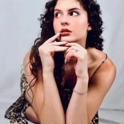 Portret kobiety z kręconymi włosami, w sukience w panterkę, z rękami blisko twarzy, z pierścionkiem i bransoletką, na białym tle. Patrzy w lewo.