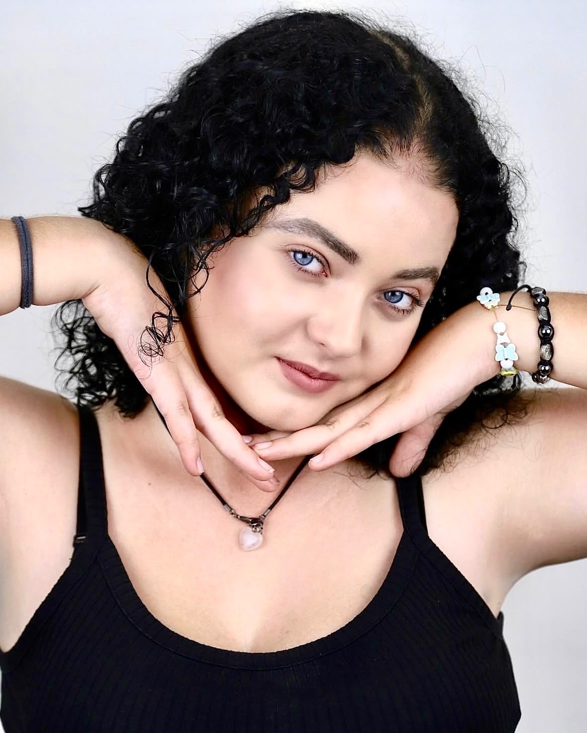 Portret kobiety z kręconymi włosami i niebieskimi oczami, pozującej z rękami przy twarzy, w czarnym topie i z biżuterią na nadgarstkach.