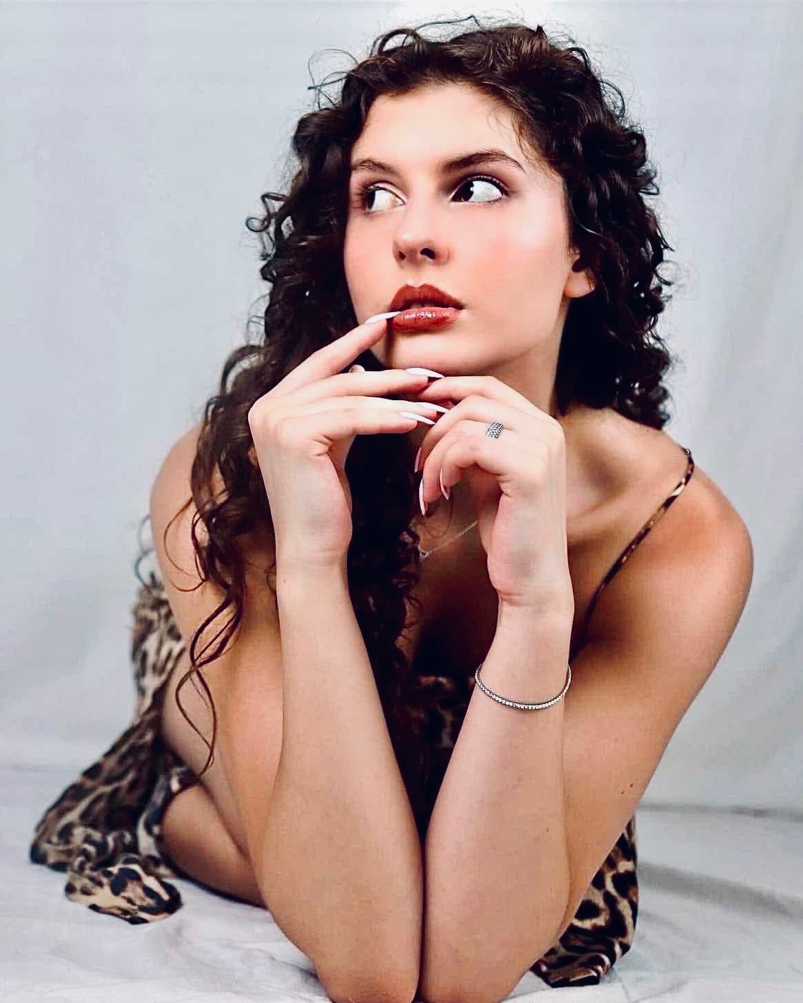 Portret kobiety z kręconymi włosami, w sukience w panterkę, z rękami blisko twarzy, z pierścionkiem i bransoletką, na białym tle. Patrzy w lewo.