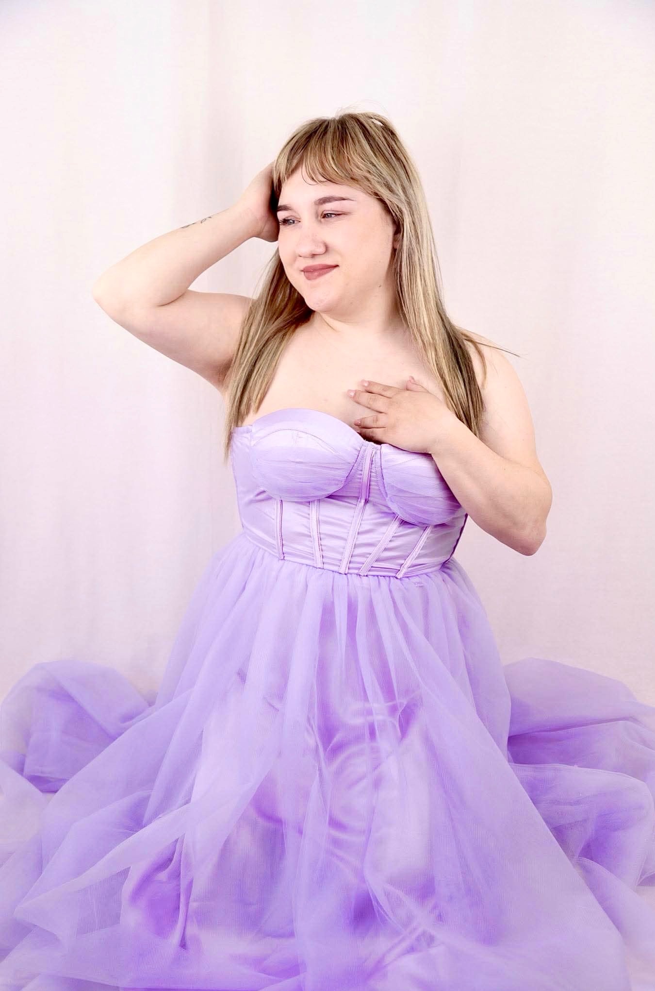 Portret kobiety w fioletowej sukni balowej, z dłonią na głowie i klatce piersiowej, na jasnym tle. Uśmiech i delikatny makijaż podkreślają naturalność.