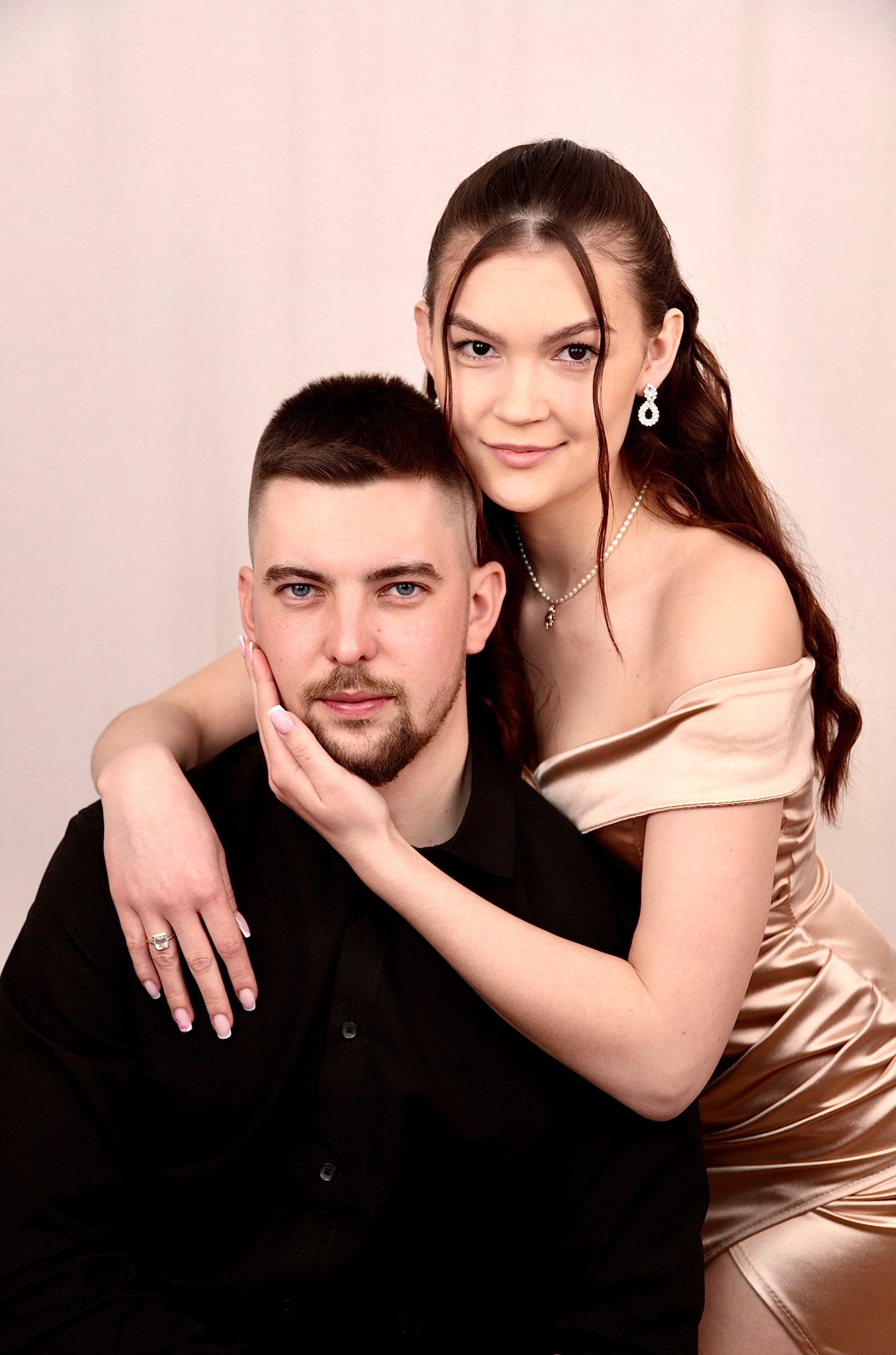 Portret pary: mężczyzna w czarnej koszuli, kobieta w złotej sukience, obejmująca go czule. Delikatny makijaż, biżuteria i jasne tło podkreślają elegancję.