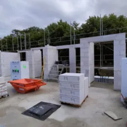 Wznoszenie ścian z bloczków betonu komórkowego na budowie domu. Widoczne rusztowania, palety z materiałem, drabina i pomarańczowa taczka budowlana. W tle zieleń drzew.