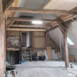 Poddasze w trakcie remontu: widoczne drewniane belki, ocieplenie ścian i fragmenty płyt karton-gips na suficie. Schody prowadzą do kolejnego pomieszczenia.