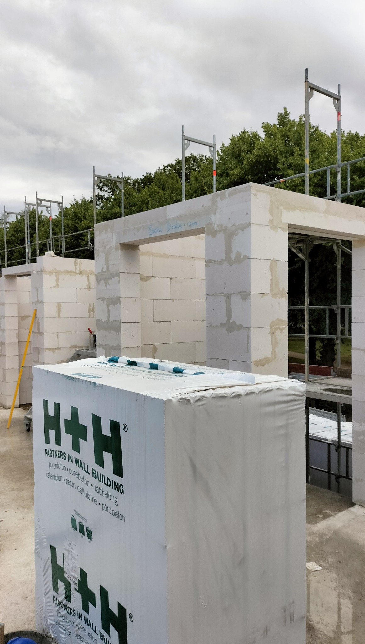 Wznoszenie ścian z białych bloczków H+H na placu budowy. Widoczne rusztowania i zapakowane bloczki. Napis 'Bad Doberan' nad otworem drzwiowym.