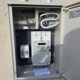 Paweł Bubeła Usługi Elektryczne - Otwarta skrzynka elektryczna na elewacji budynku, widoczne bezpieczniki, licznik energii i przewody. Biała kartka z numerem 1580 wewnątrz.