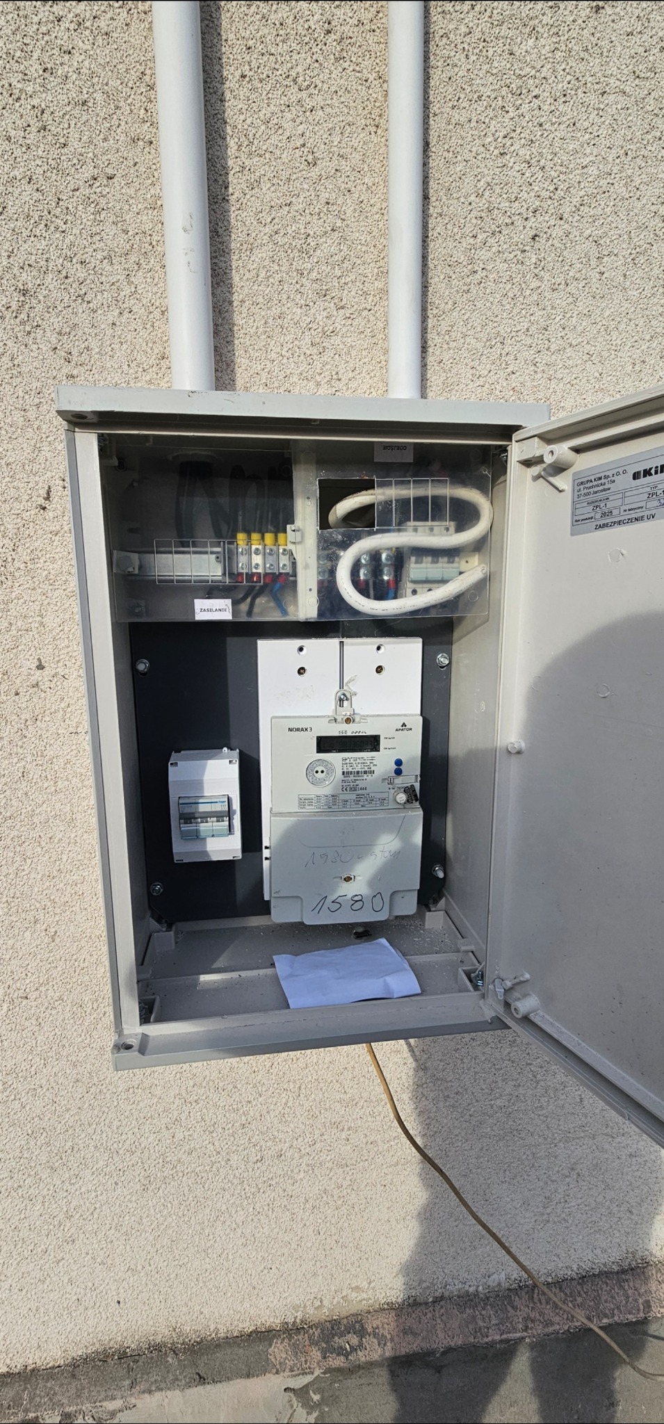 Otwarta skrzynka elektryczna na elewacji budynku, widoczne bezpieczniki, licznik energii i przewody. Biała kartka z numerem 1580 wewnątrz.