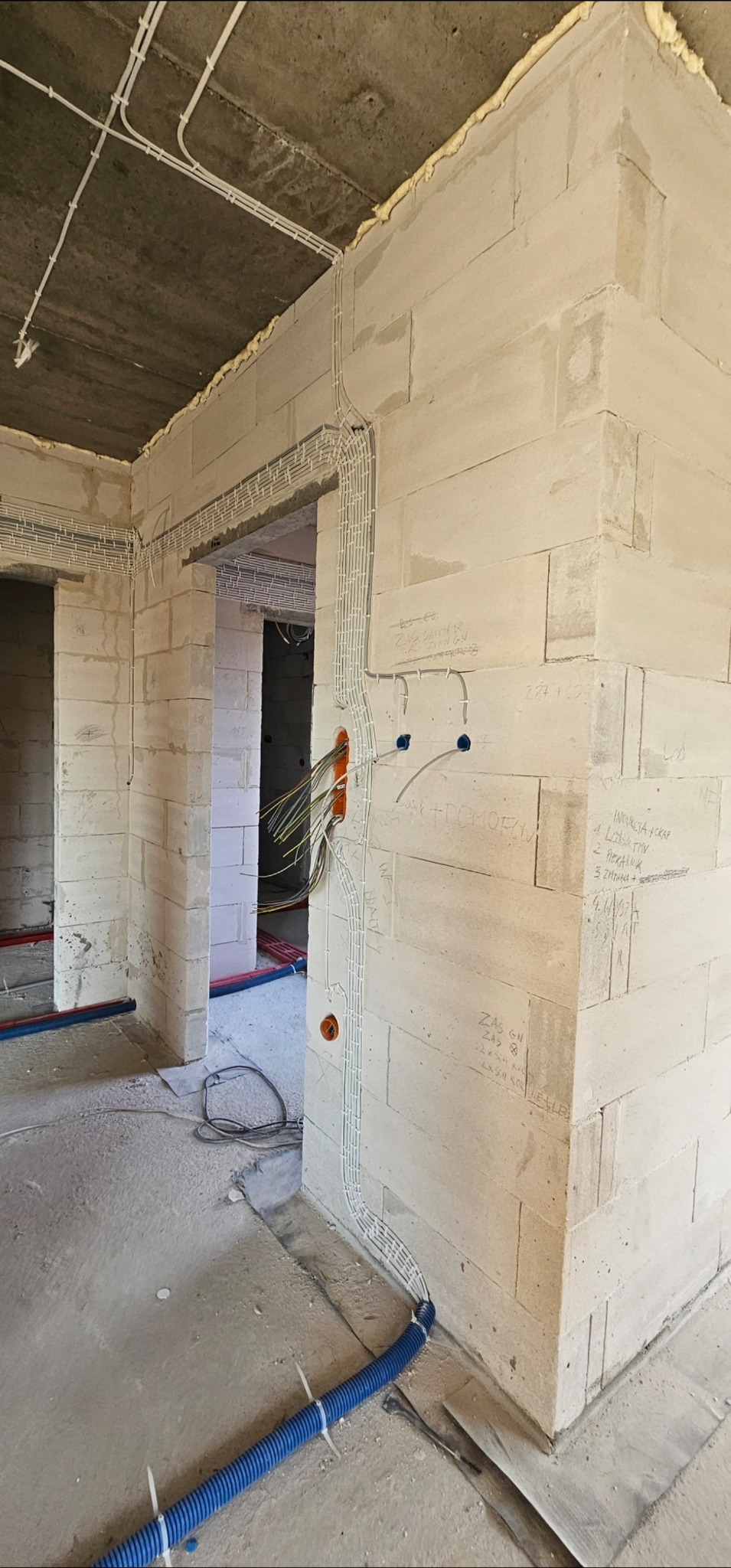Surowy stan: białe ściany z betonu komórkowego, widoczne kable w korytkach i peszlach, instalacja elektryczna w trakcie montażu, otwory pod gniazdka.