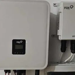 Biała obudowa inwertera fotowoltaicznego FOX ESS z wyświetlaczem i skrzynka EPS BOX-TP zamontowane na ścianie, widoczne przewody i elementy instalacji.