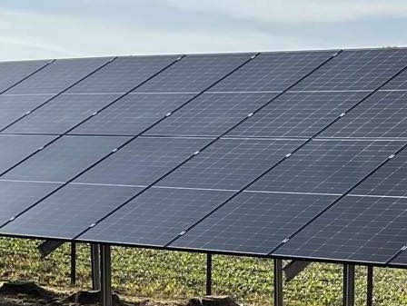 Rząd paneli fotowoltaicznych na metalowej konstrukcji, na tle nieba i zieleni. Widok z dołu, ukazujący strukturę i montaż paneli.