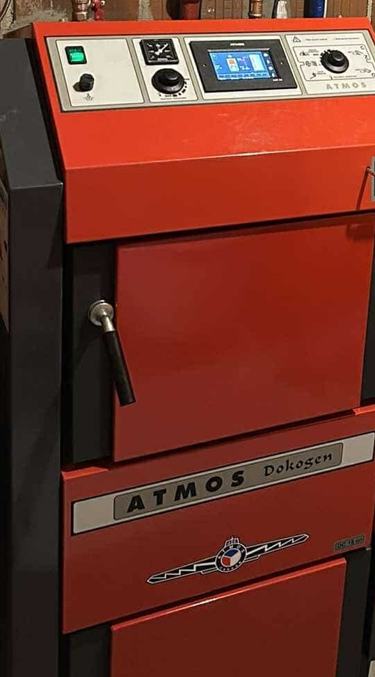 Czerwony piec CO Atmos Dokogen z czarną klamką i panelem sterowania. Widoczny fragment ściany. Model z wyświetlaczem, przyciskiem i pokrętłami.