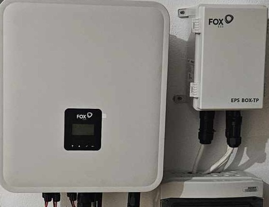 Biała obudowa inwertera fotowoltaicznego FOX ESS z wyświetlaczem i skrzynka EPS BOX-TP zamontowane na ścianie, widoczne przewody i elementy instalacji.