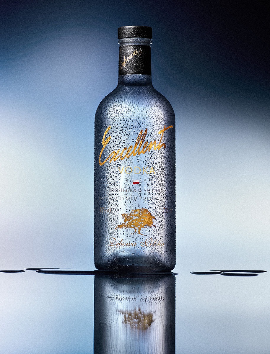 Wysokiej jakości fotografia produktowa: butelka wódki z kroplami wody i złotymi napisami na tle gradientu niebieskiego, z odbiciem na powierzchni.
