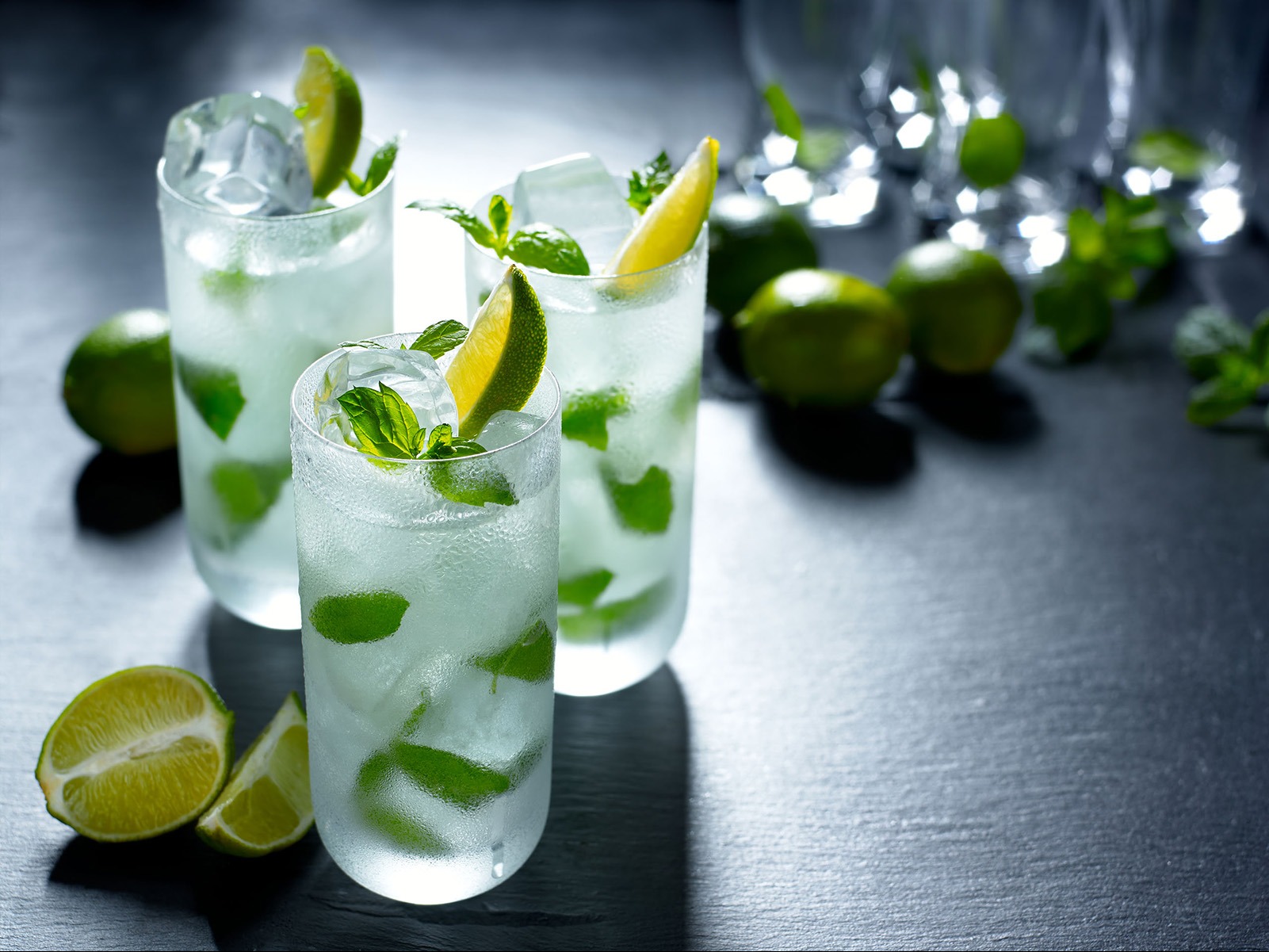 Trzy szklanki orzeźwiającego mojito z lodem, limonką i miętą na ciemnym tle. W tle rozmyte owoce limonki i dodatkowe szklanki. Chłodny, letni napój.