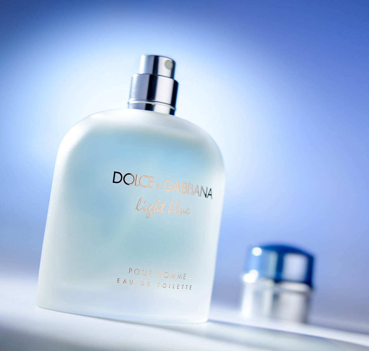 Flakon męskich perfum Dolce & Gabbana Light Blue na jasnoniebieskim tle. Srebrny atomizer i napis w kolorze złota. Korek flakonu poza ostrością.