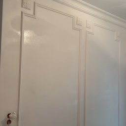 SV REM-BUD - Biała ściana z dekoracyjnymi listwami i gładzią, widoczne puszki elektryczne bez osprzętu. Wykończenie w stylu klasycznym.