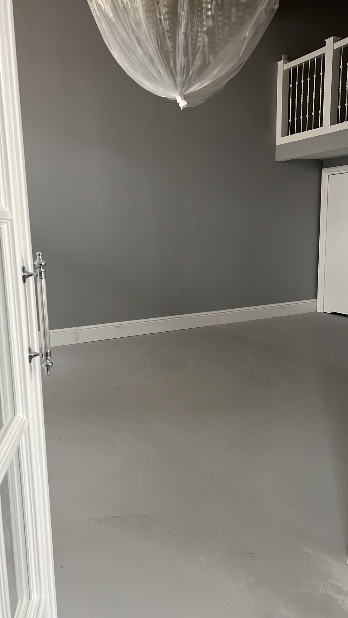 Szara ściana po malowaniu, zabezpieczony żyrandol folią, widoczny fragment balustrady i drzwi. Podłoga szpachlowana, przygotowana do dalszych prac wykończeniowych.