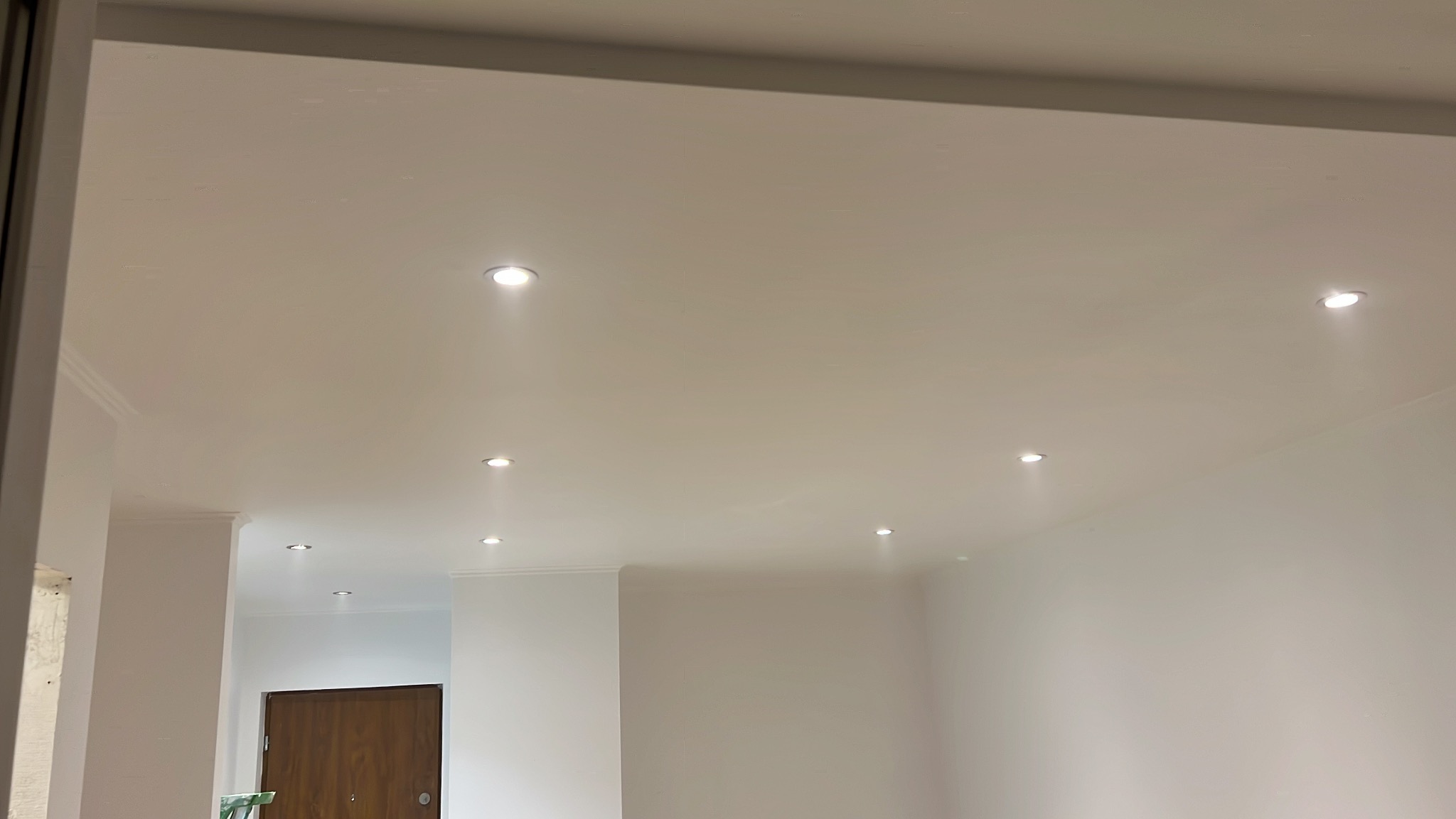 Białe wnętrze z nowo zamontowanymi, punktowymi lampami sufitowymi, widok z dołu. Listwy dekoracyjne przy suficie. W tle widoczny fragment drzwi.