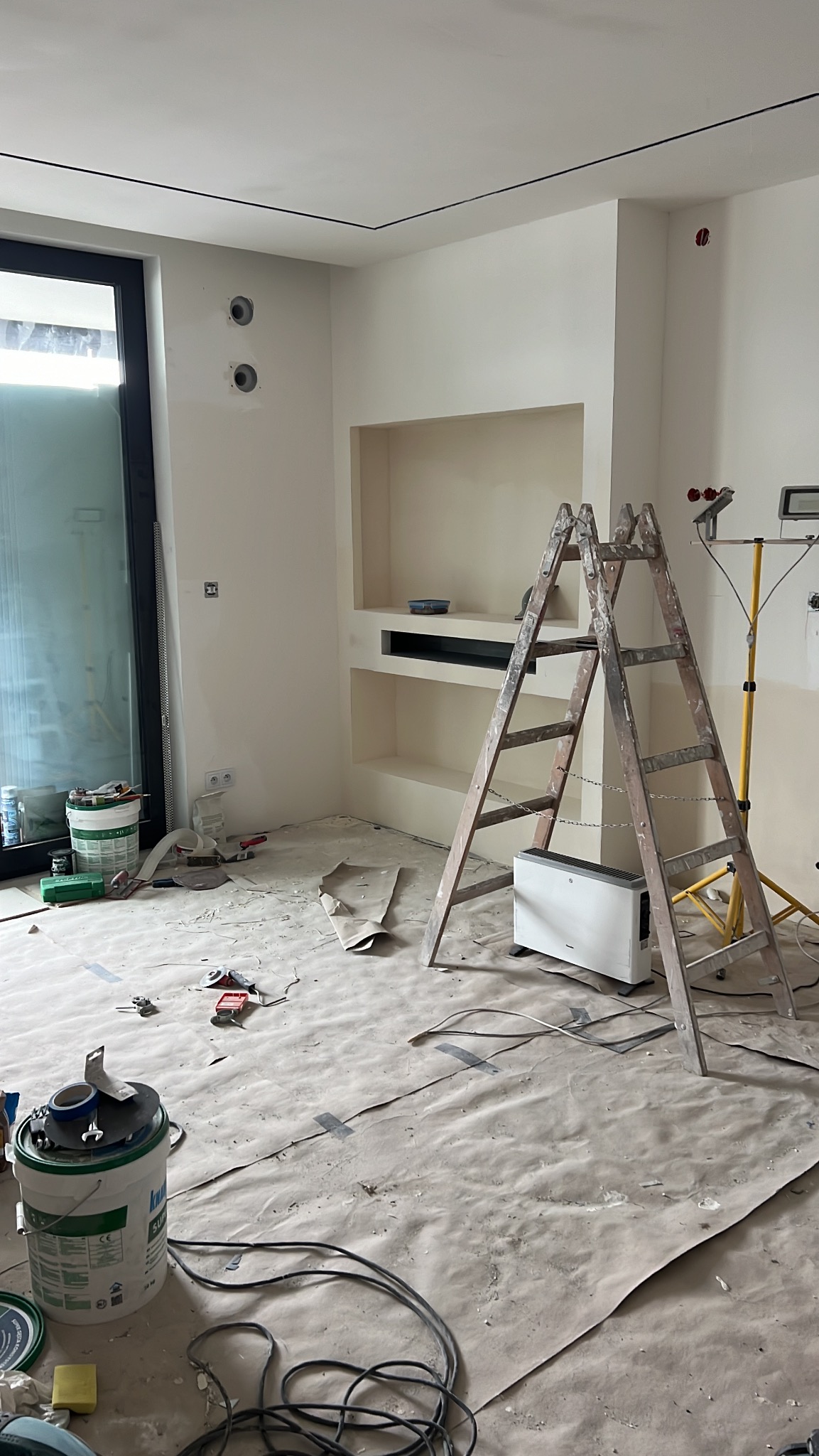 Pokój w trakcie malowania na kremowo. Widoczna drabina, wiadro z farbą, zakryta podłoga, wnęka w ścianie i lampa halogenowa. Etap prac wykończeniowych.