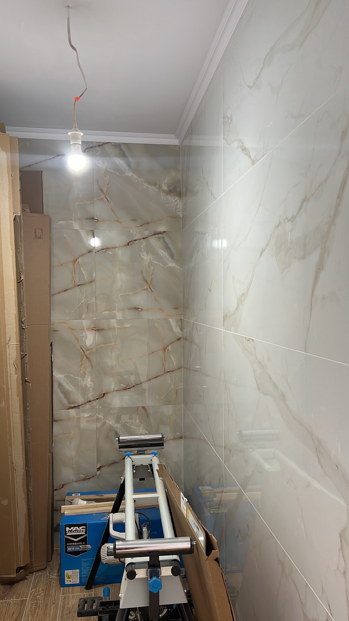 Wykończona ściana łazienki z marmurowymi płytkami, widoczny fragment narzędzia do cięcia płytek i kartony. Oświetlenie sufitowe z żarówką.
