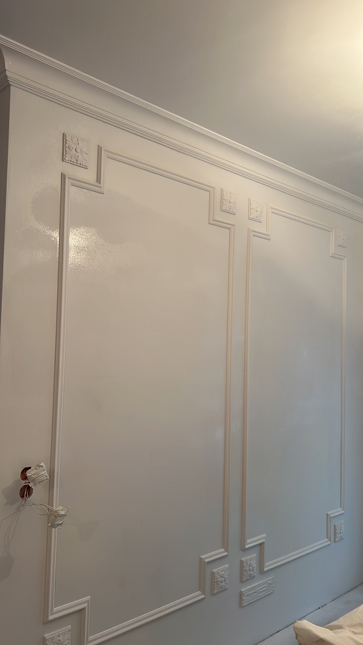 Biała ściana z dekoracyjnymi listwami i gładzią, widoczne puszki elektryczne bez osprzętu. Wykończenie w stylu klasycznym.