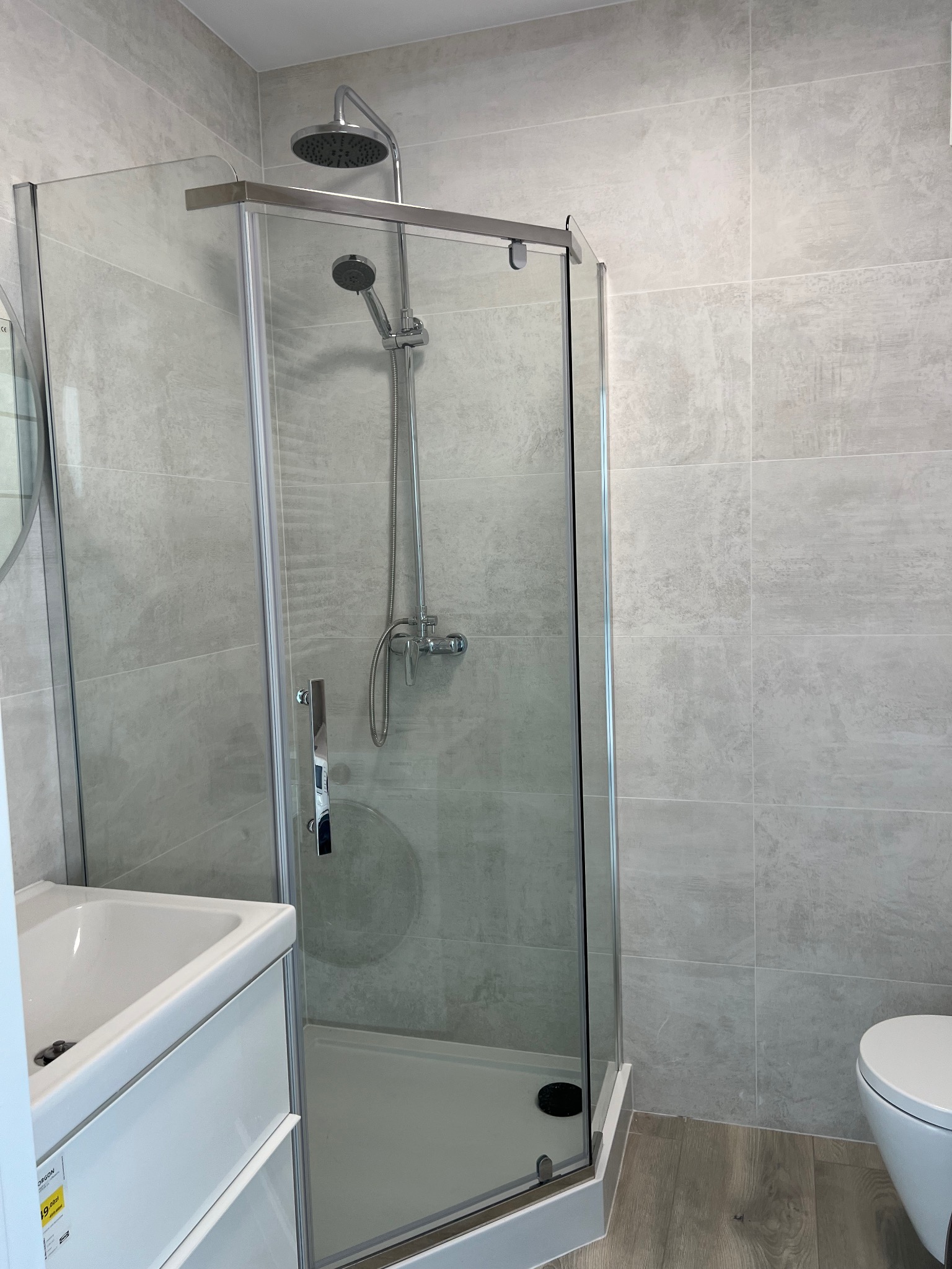 Jasna łazienka z kabiną prysznicową typu walk-in, szare płytki na ścianach i drewnopodobna podłoga. Umywalka z szafką i toaleta w tle.