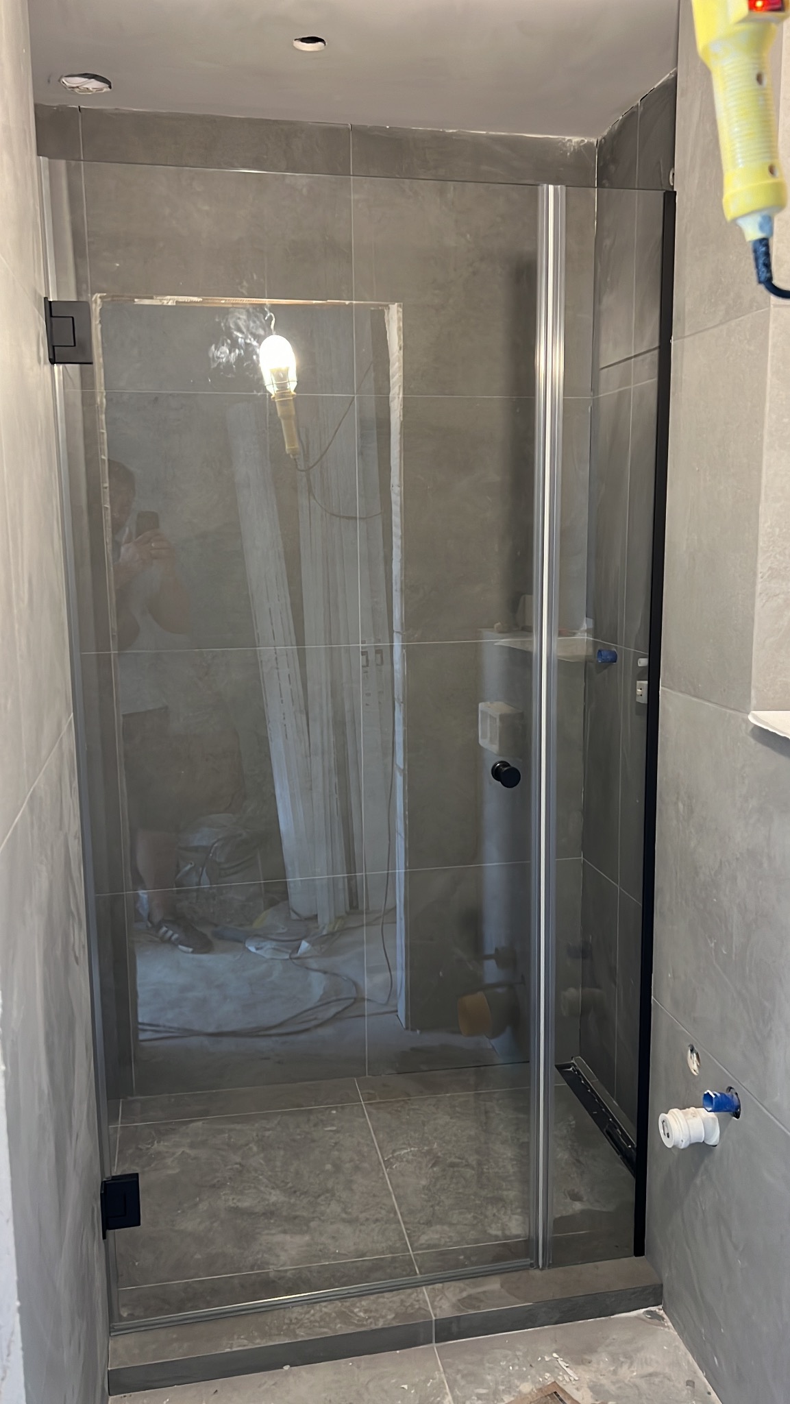 Szklana kabina prysznicowa z czarnymi okuciami w szarej łazience w trakcie wykańczania, widoczne narzędzia i lampa robocza. Minimalistyczny design.