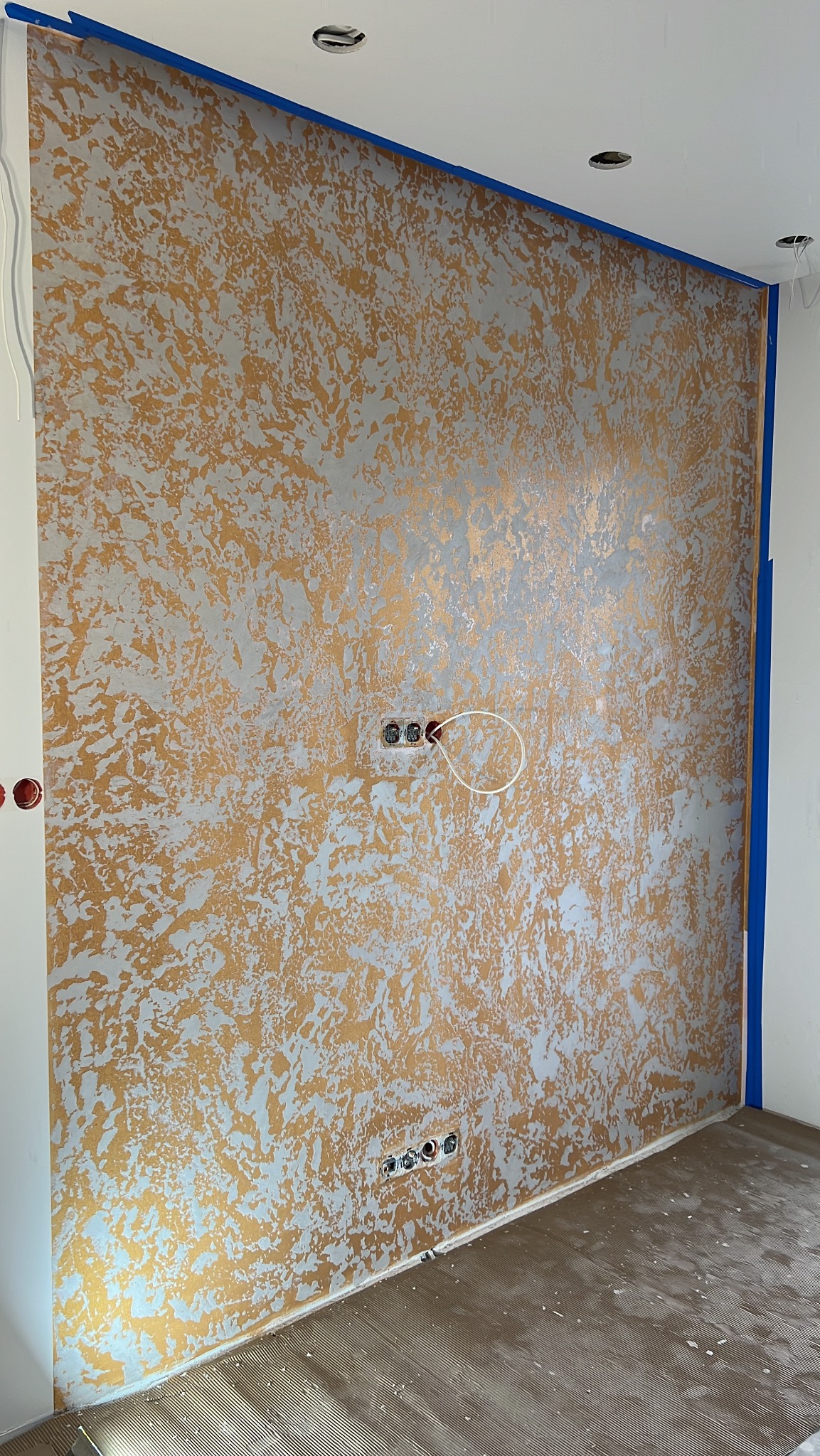 Dekoracyjna ściana w odcieniach złota i srebra z widocznymi gniazdkami elektrycznymi, zabezpieczona niebieską taśmą malarską przy suficie i ścianach.