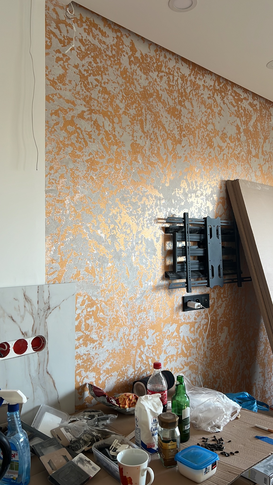 Ściana z dekoracyjnym tynkiem w odcieniach złota i szarości, z zamontowanym uchwytem TV. Na pierwszym planie widoczne akcesoria i materiały remontowe.