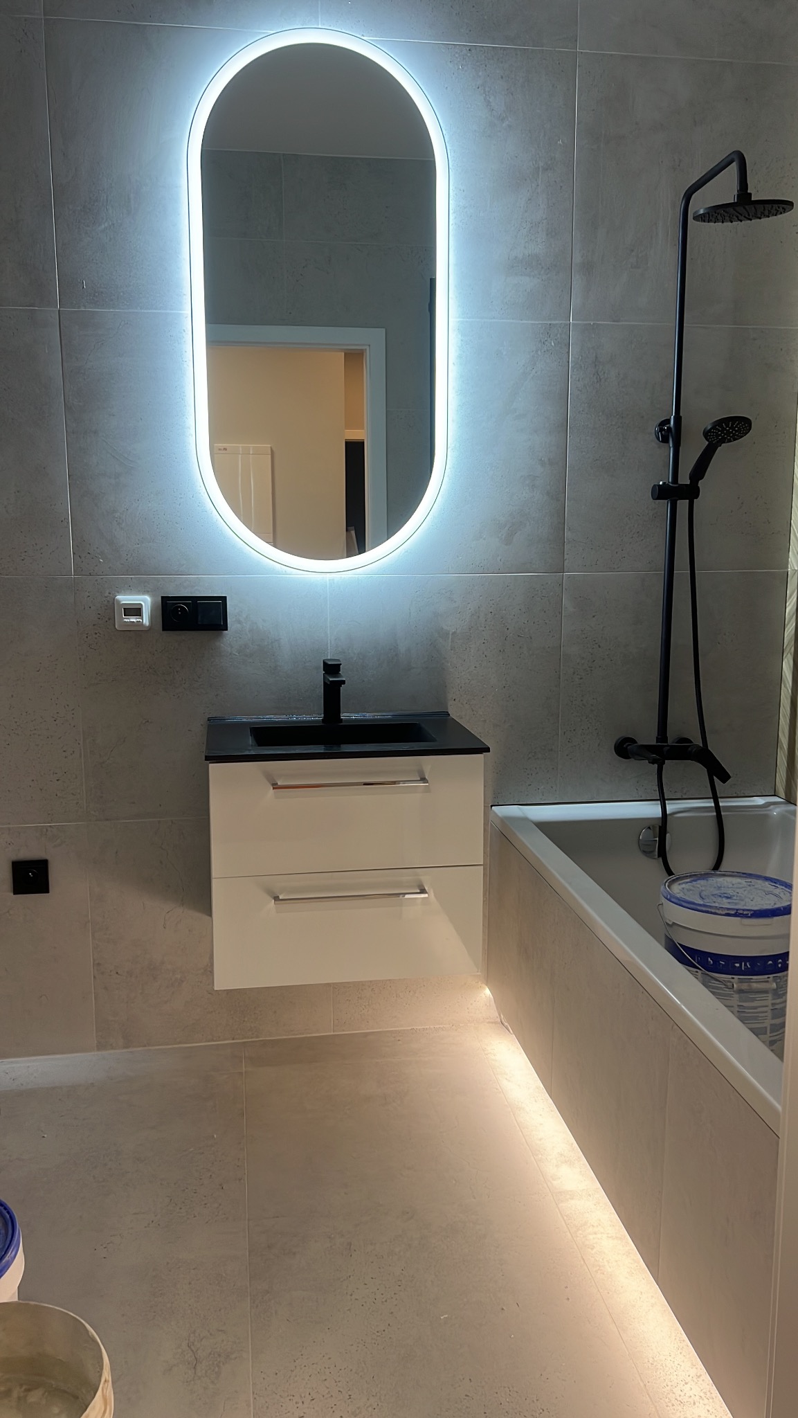 Nowoczesna łazienka z owalnym lustrem LED, czarną armaturą i podświetleniem pod szafką umywalkową. Szare płytki na ścianach i podłodze.