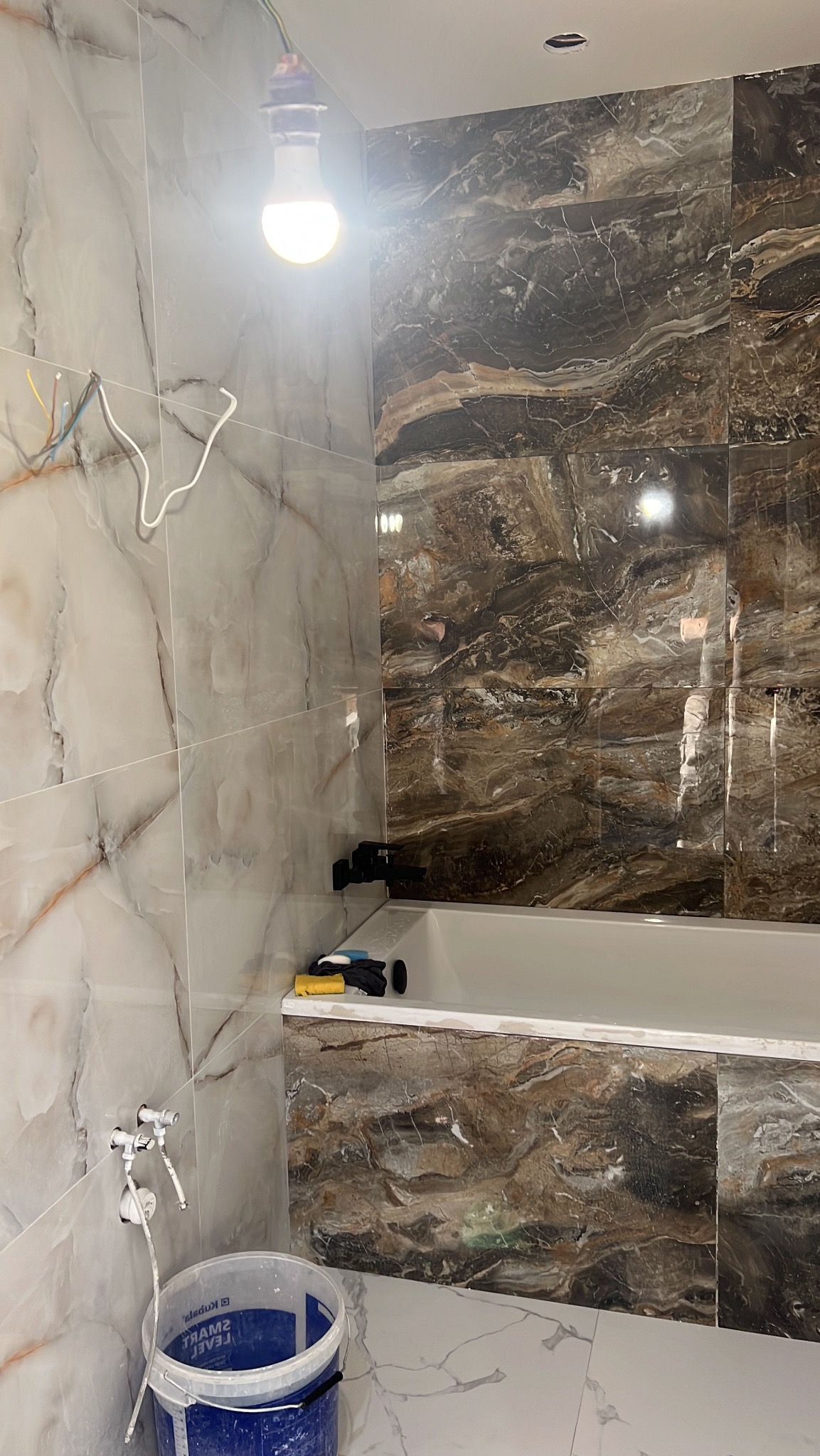 Wykończona ściana z marmurowych płytek nad wanną w łazience. Widoczny kubeł, narzędzia i prowizoryczne oświetlenie. Płytki w odcieniach bieli i brązu.