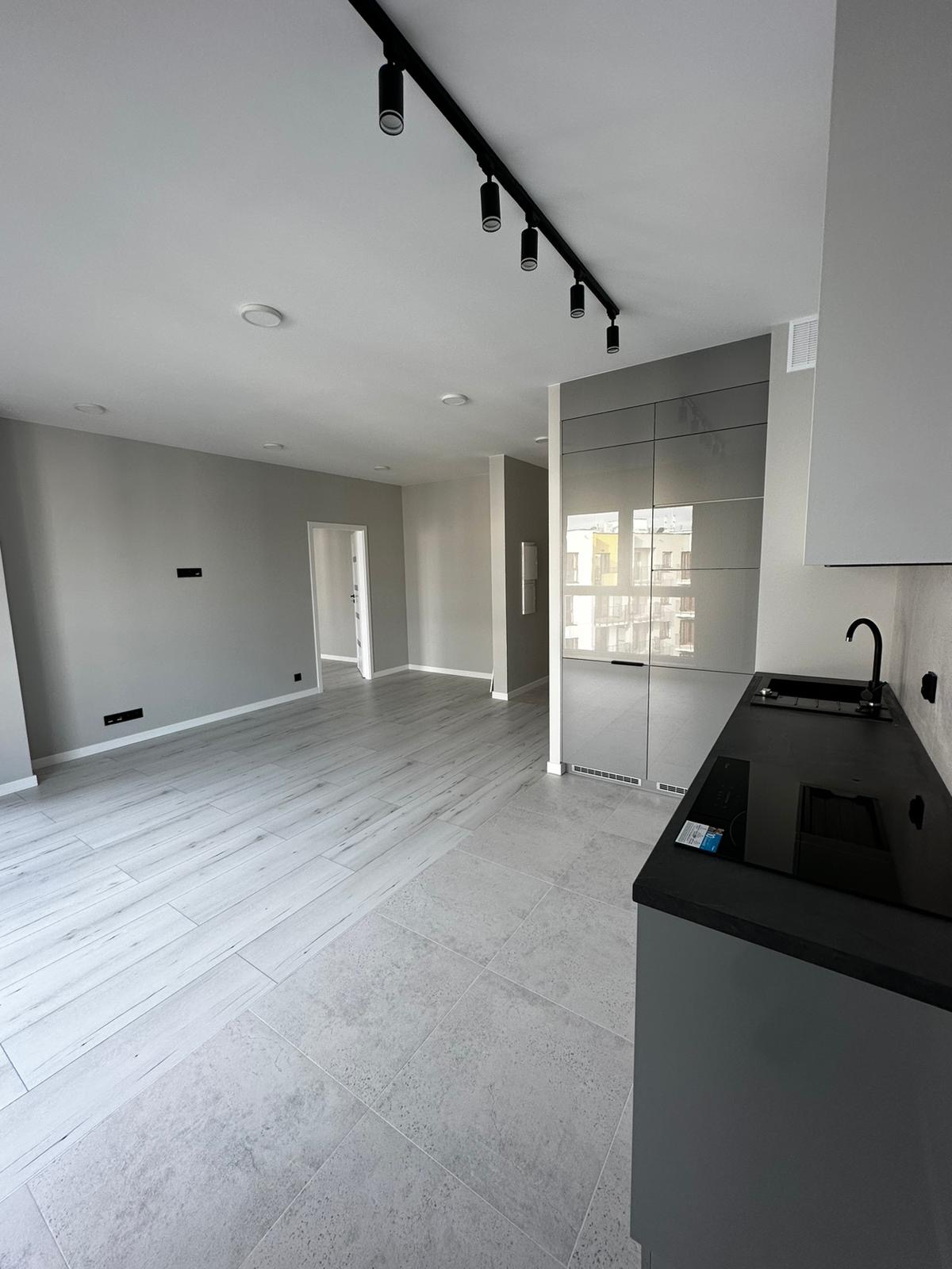 Jasne, nowoczesne wnętrze po remoncie: szare ściany, panele podłogowe imitujące drewno, płytki w kuchni, czarne oświetlenie szynowe i zabudowa kuchenna.