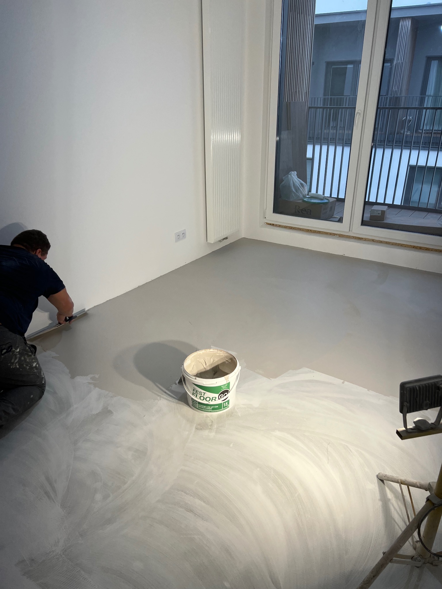 Aplikacja mikrocementu na podłodze w jasnym wnętrzu. Widoczny pracownik rozprowadzający masę, wiadro z produktem 'Fest Floor' i lampa oświetleniowa.