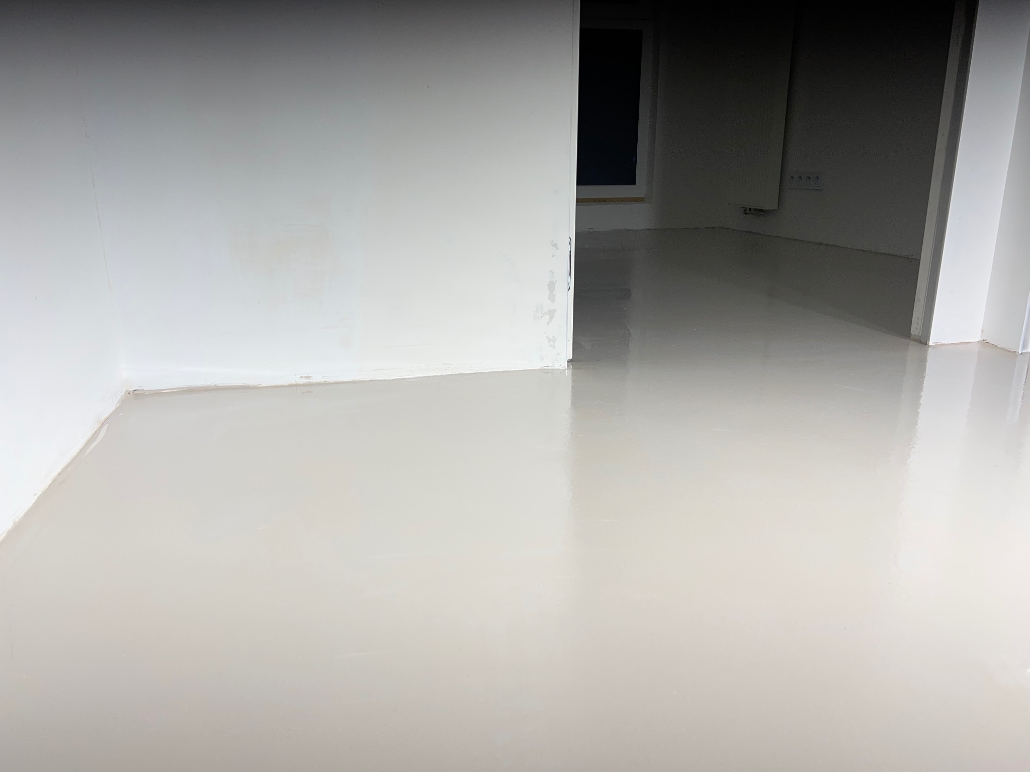 Wnętrze po wylaniu samopoziomującej wylewki na podłodze. Widoczne gładkie, jasne podłoże oraz fragment ściany z oknem w tle. Minimalistyczny efekt.