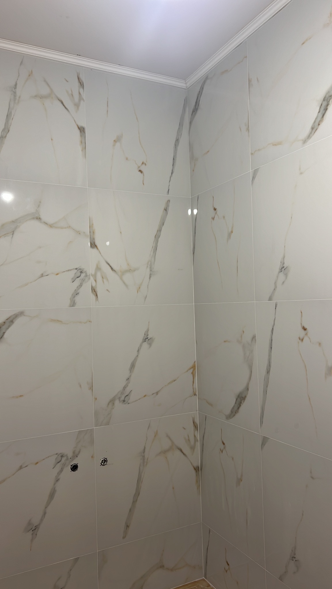 Wykończona ściana w łazience z białych, marmurkowych płytek, z widocznymi otworami na armaturę i listwą dekoracyjną przy suficie. Estetyczne połączenie płytek w rogu.