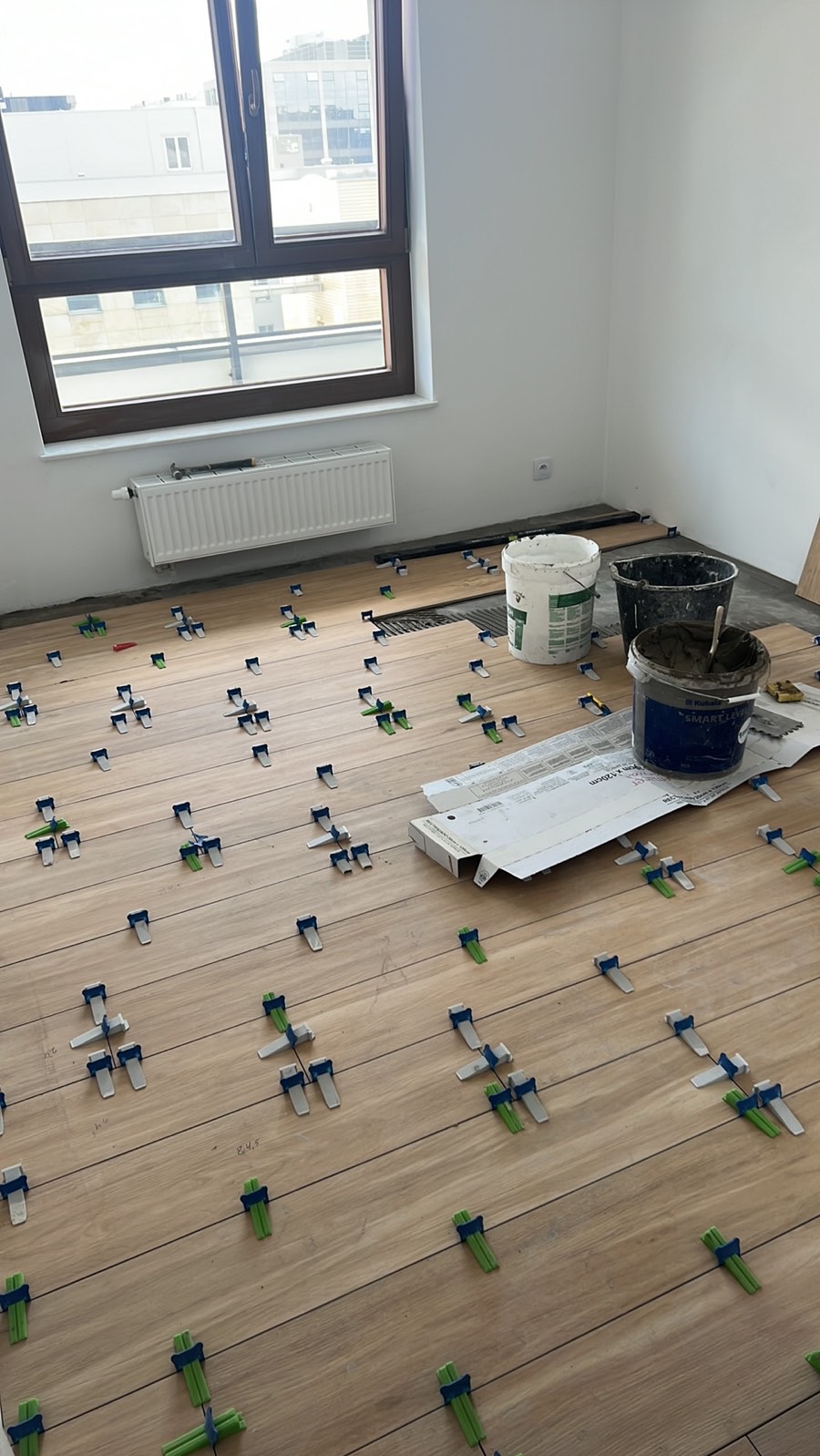 Układanie drewnopodobnych płytek z użyciem klipsów poziomujących w Ząbkach. Widoczne wiadra z zaprawą i okno z widokiem na miasto.